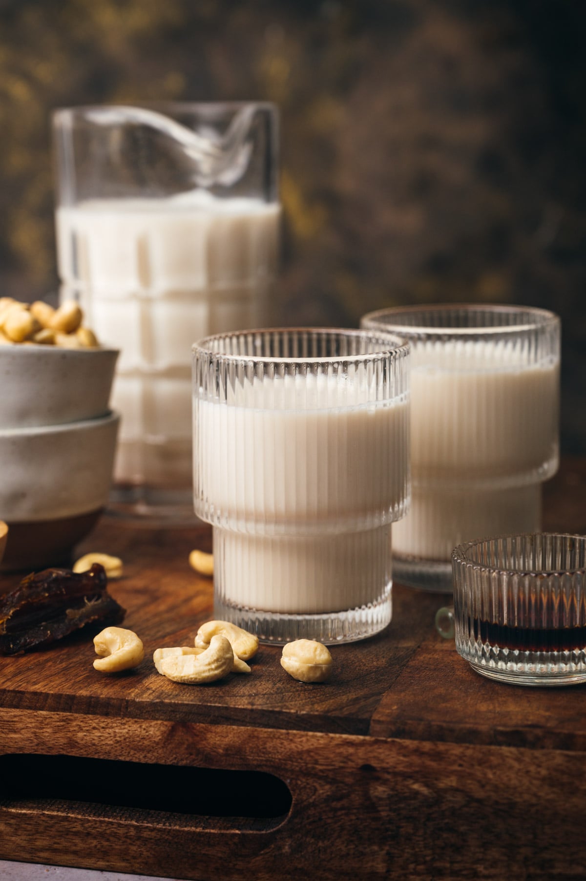 Two glasses of cashew milk on a wooden board, surrounded by cashew nuts and a partial view of a larger glass jug in the background.