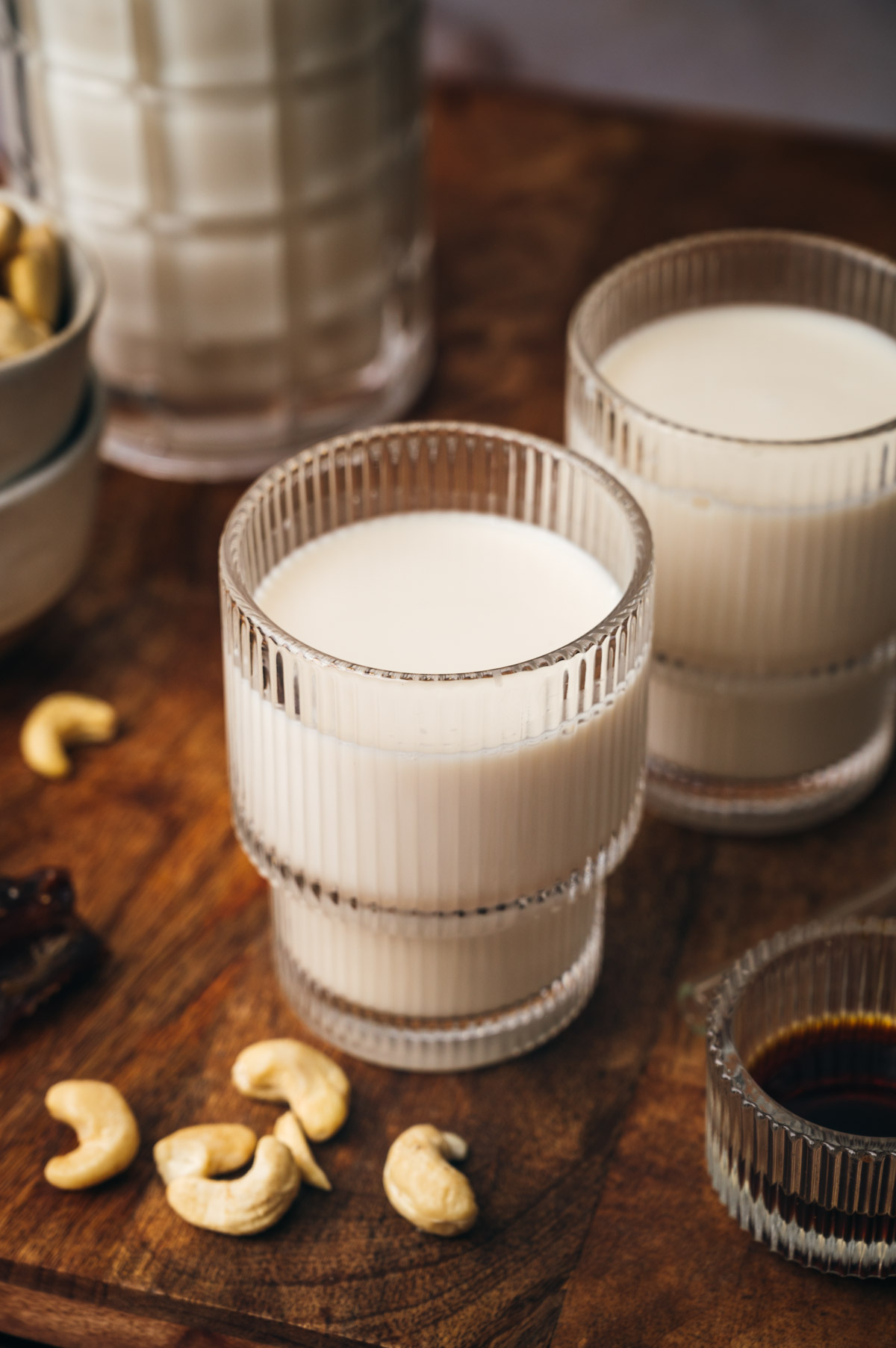 Two glasses of milk on a wooden surface surrounded by cashew nuts and a small bowl of syrup. Other ingredients and a pitcher are in the background.