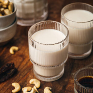 Three glasses of cashew milk on a wooden table, surrounded by a few whole cashews and other small dishes.