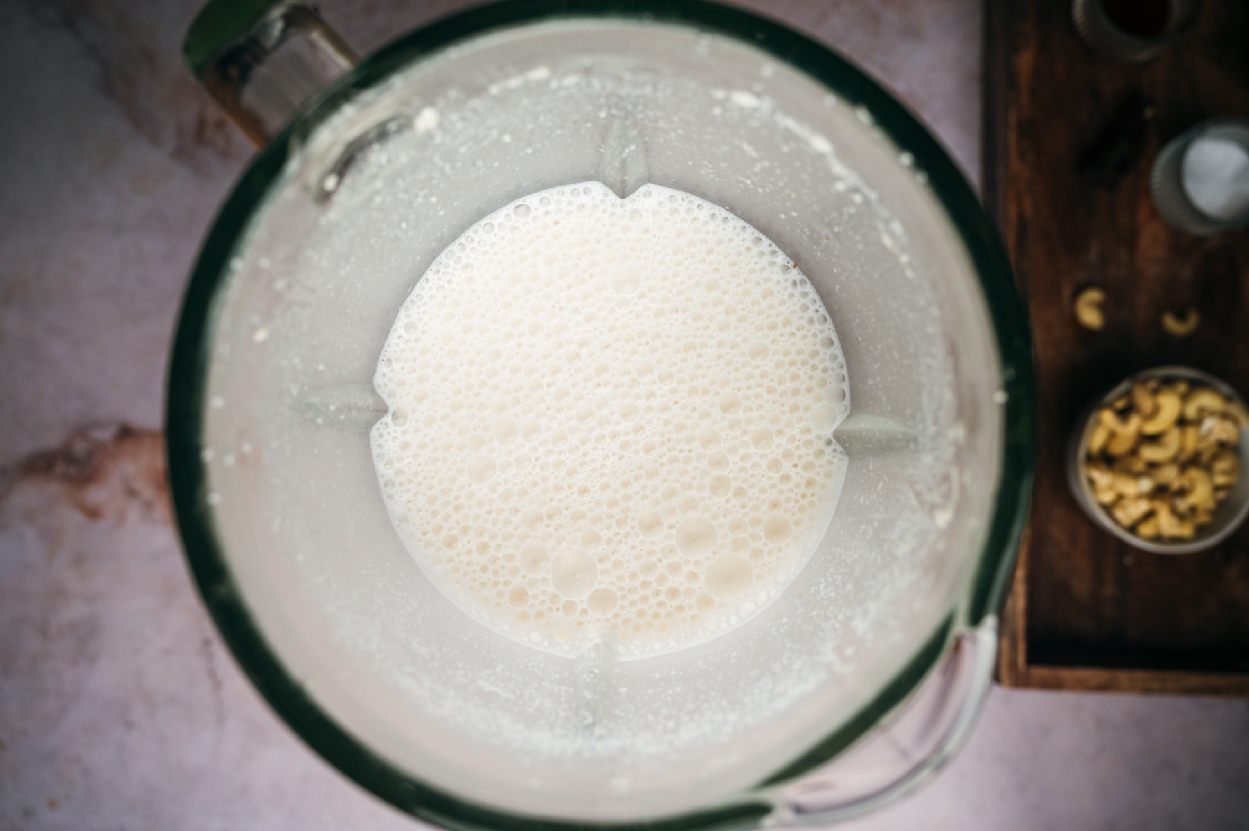 Top view of a blender filled with frothy liquid, likely a smoothie or juice blend, placed on a countertop with some garnish ingredients visible in the background.