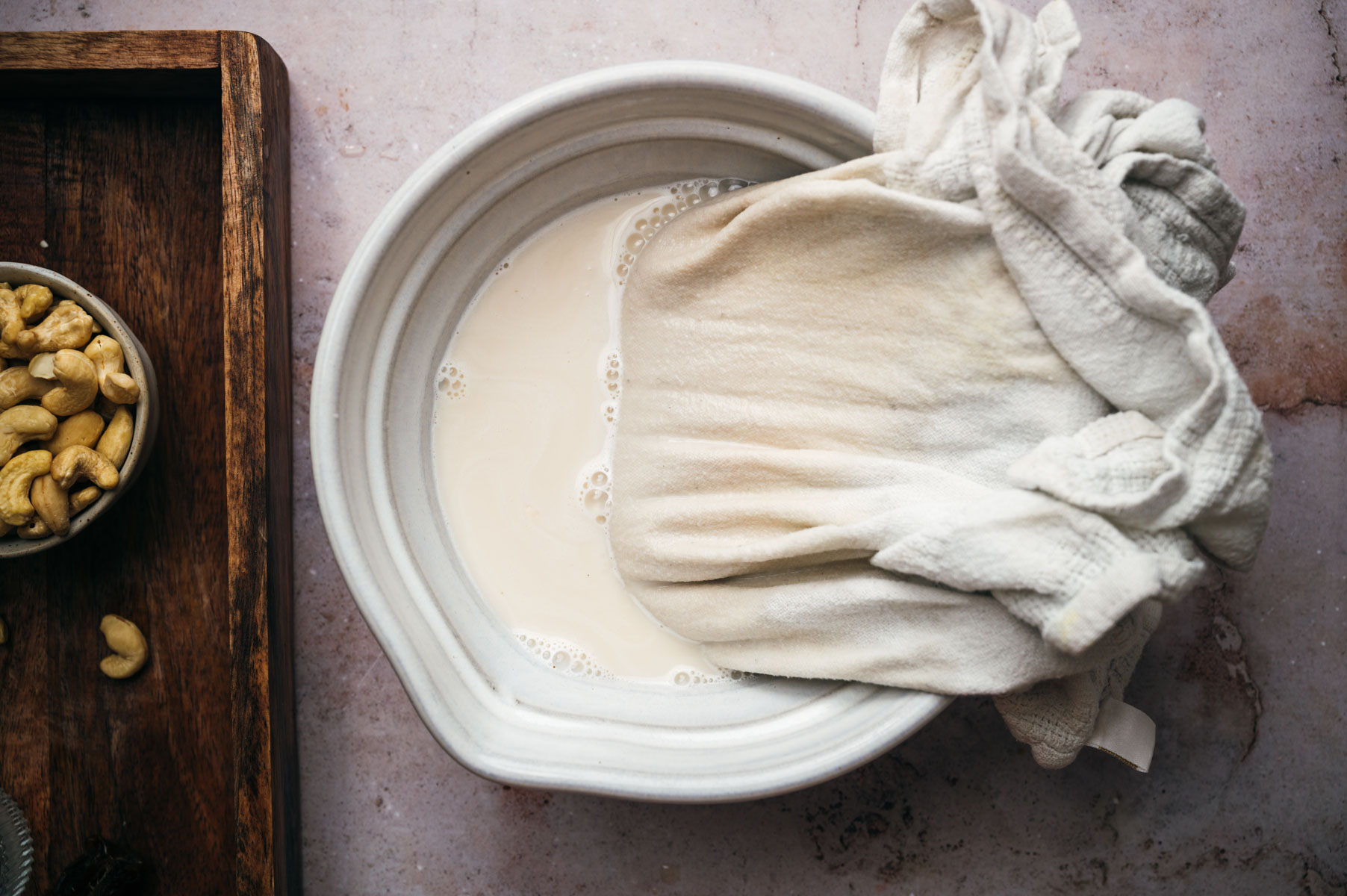 A bowl of cashew milk being strained through a cloth, with a small bowl of whole cashews on the left side of the image.