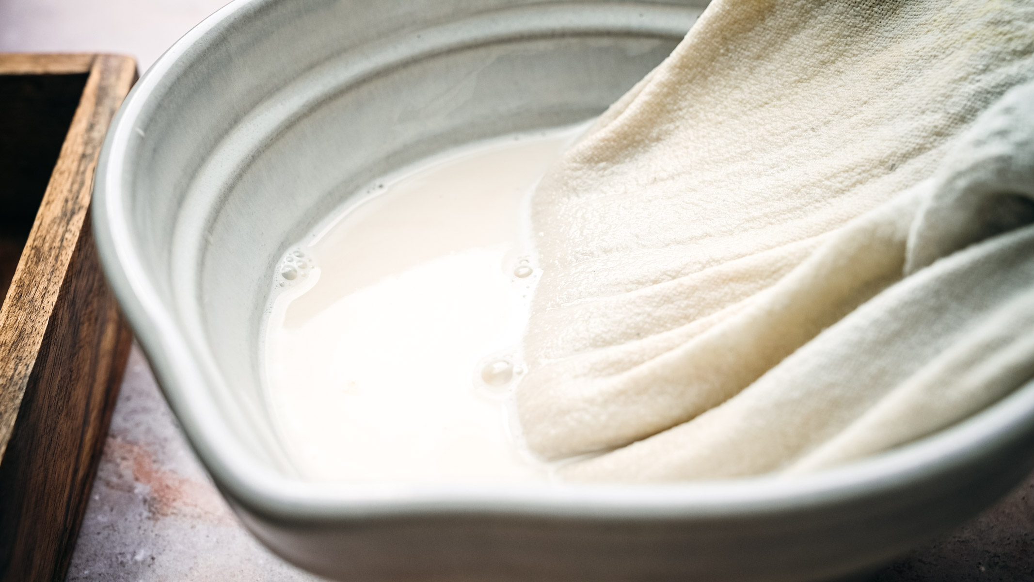 A close-up of a bowl containing a white liquid, with a cloth partially submerged, appearing to strain the liquid.