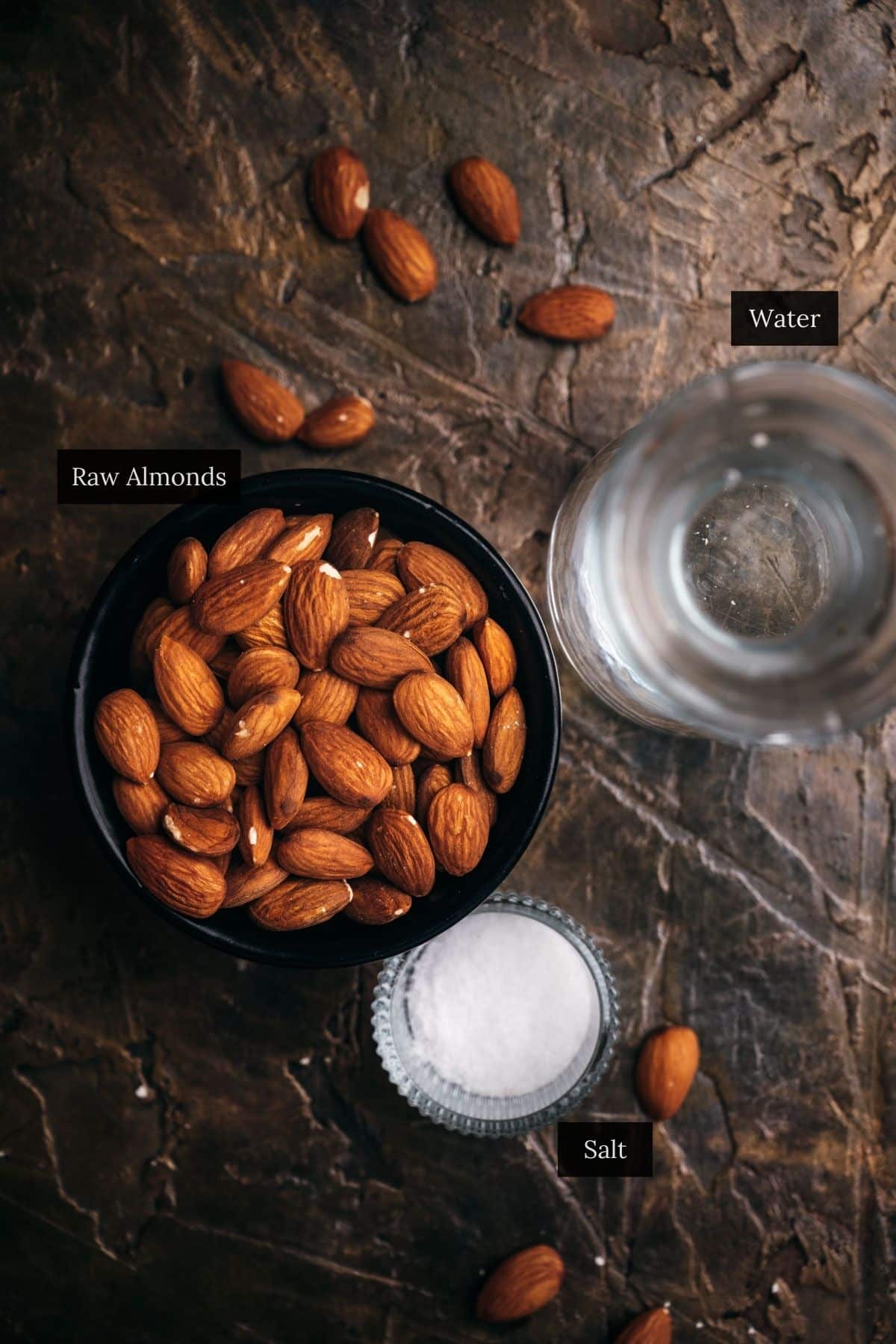 A bowl of raw almonds, a small glass bowl of salt, and a glass of water are arranged on a textured surface.