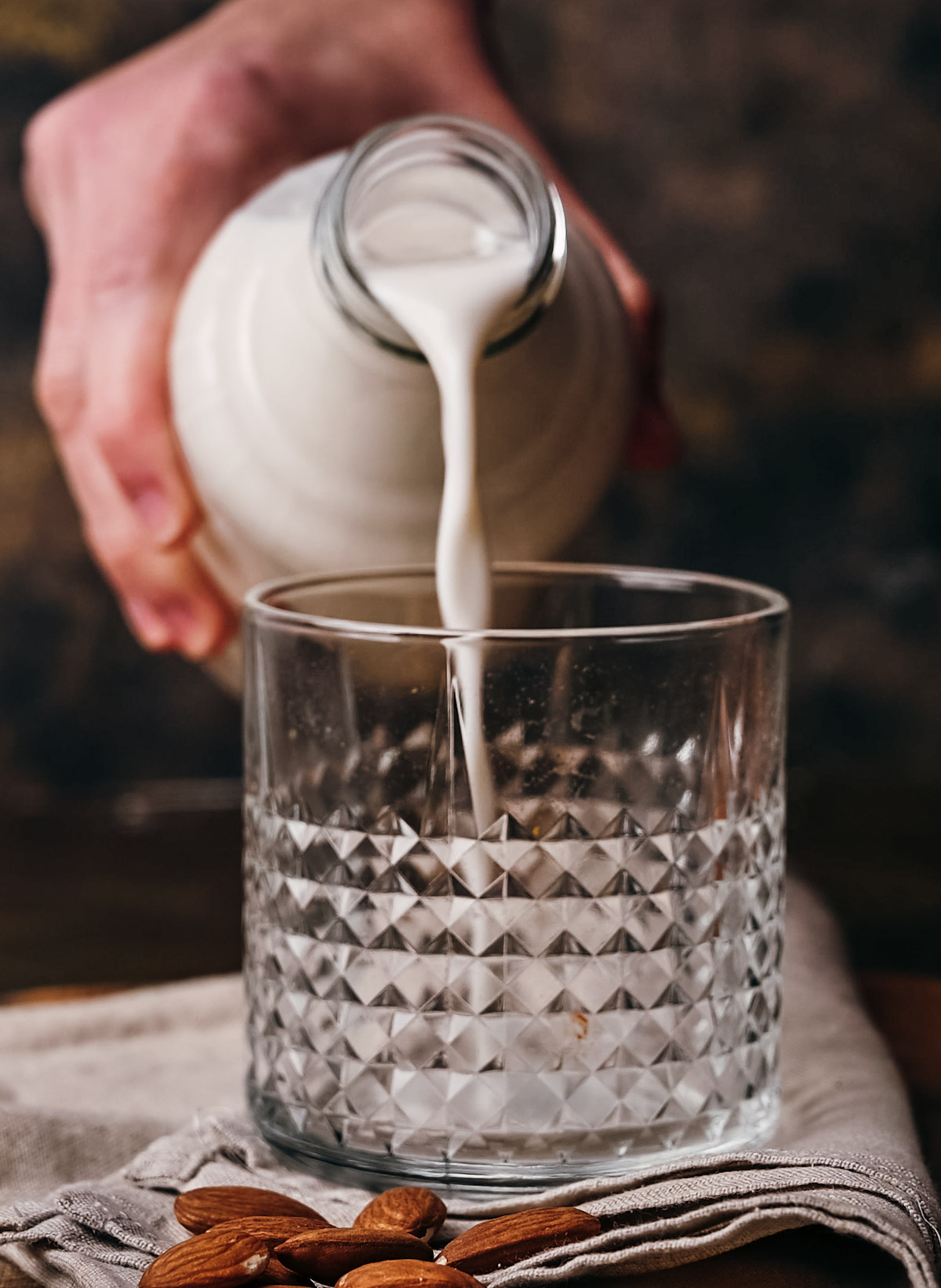 A person pours milk from a ceramic jug into a textured glass on a table with almonds scattered around.