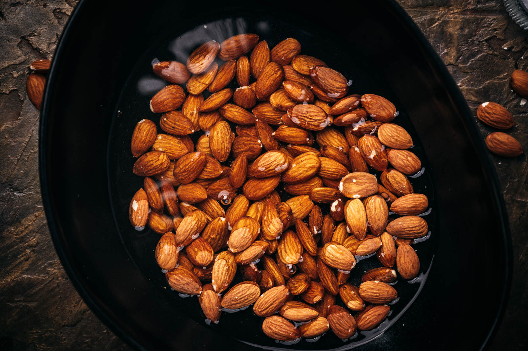 Almonds soaking in water in a black bowl on a textured stone surface.