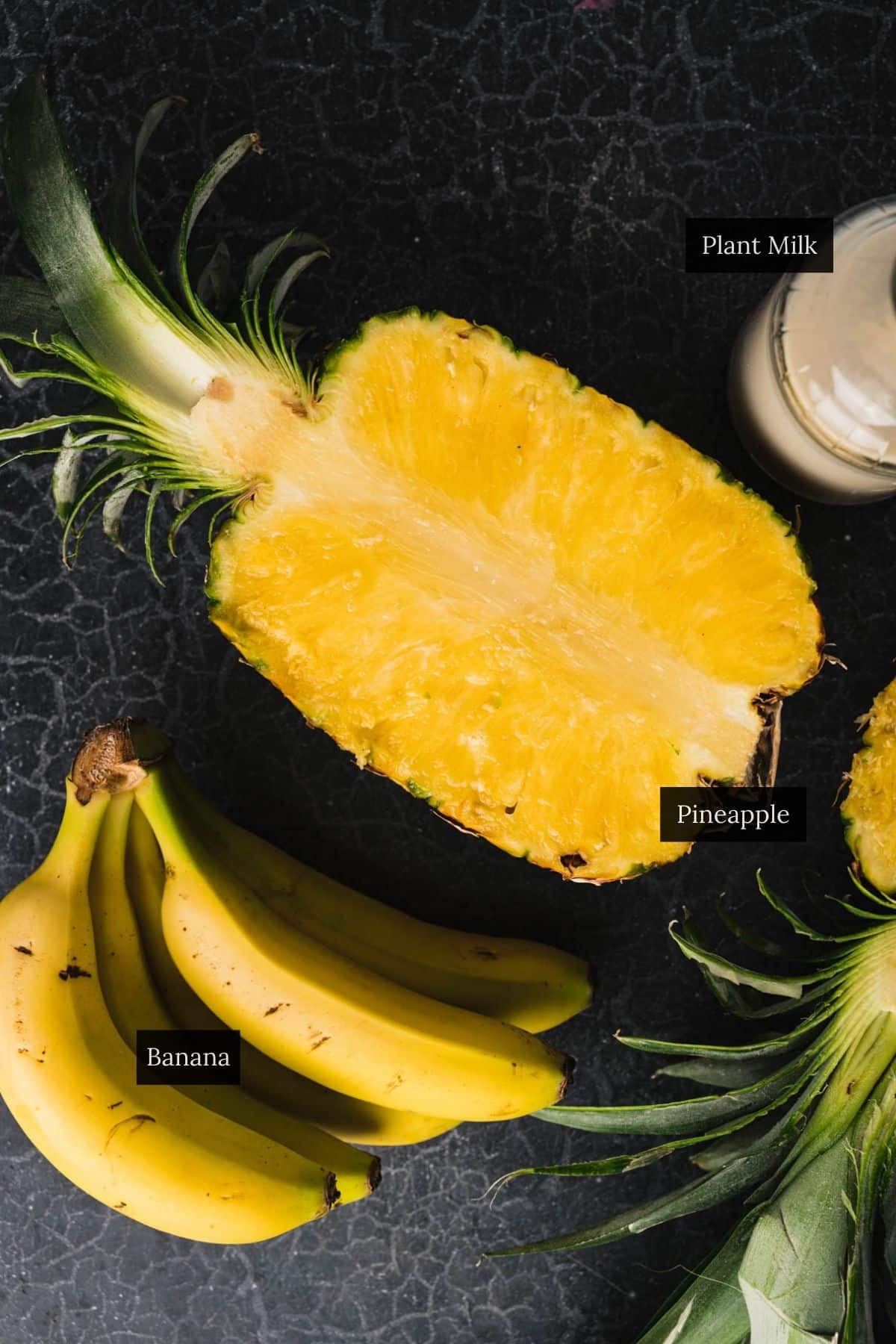 A halved pineapple, a bunch of bananas, and a bottle of a creamy liquid are placed on a dark, textured surface.
