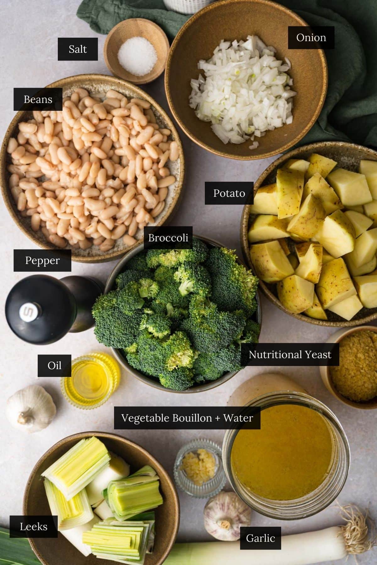 Overhead view of various ingredients in bowls, labeled as salt, onion, diced potato, nutritional yeast, chopped broccoli for vegan broccoli soup.