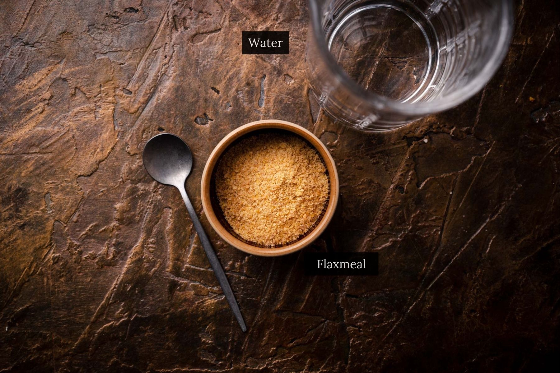 A glass of water, a bowl of flaxmeal for making a flax egg, and a spoon are arranged on a textured brown surface.
