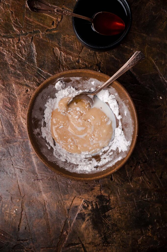 A bowl containing tahini and flour with a spoon on a rustic table, accompanied by a small dish of syrup.