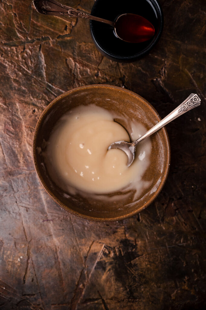 A bowl of porridge with a spoon, accompanied by a small bowl of syrup on a rustic wooden table.