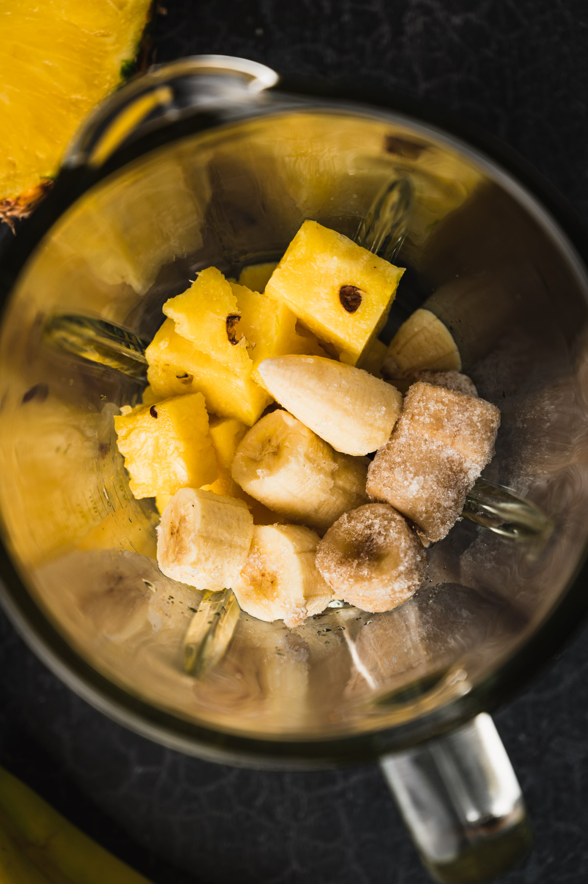 A blender jar containing chunks of pineapple, banana, and ice placed on a dark surface.