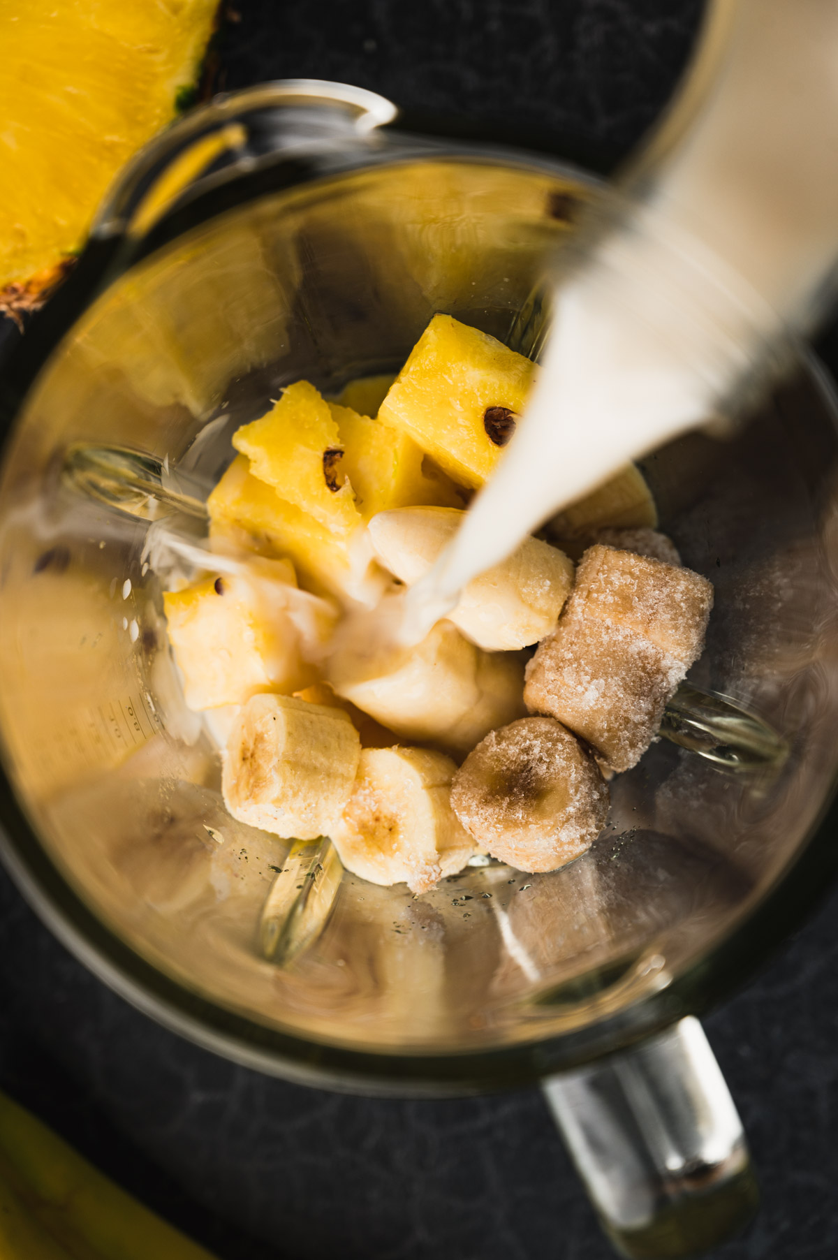 A blender filled with banana slices, pineapple chunks, and sugar being topped with a pouring liquid, possibly milk, on a dark surface.
