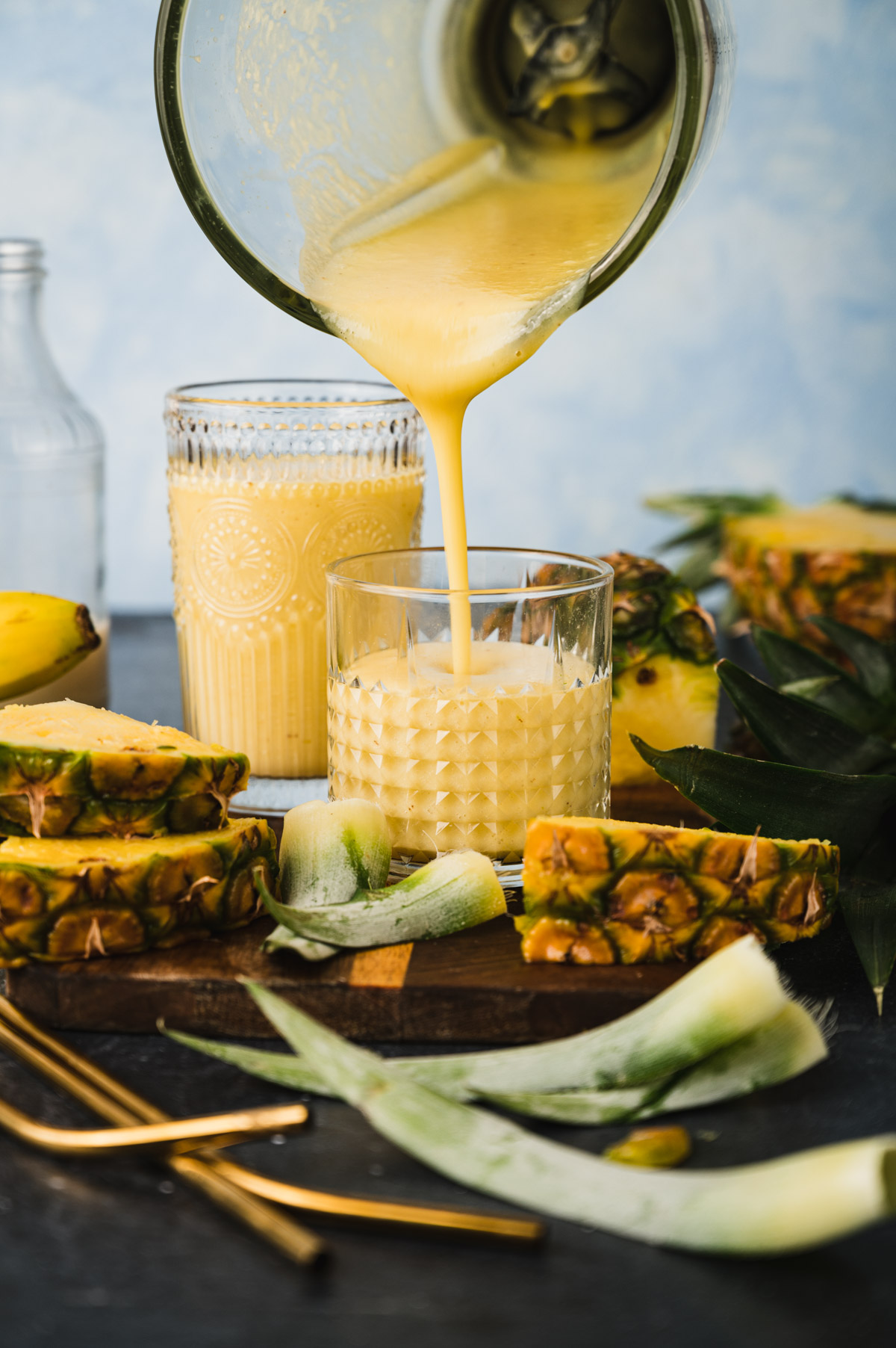 A blender pours a yellow smoothie into a glass. Another glass of smoothie and several pineapple slices are on a dark surface. There are also banana pieces, green leaves, and metal straws in the scene.