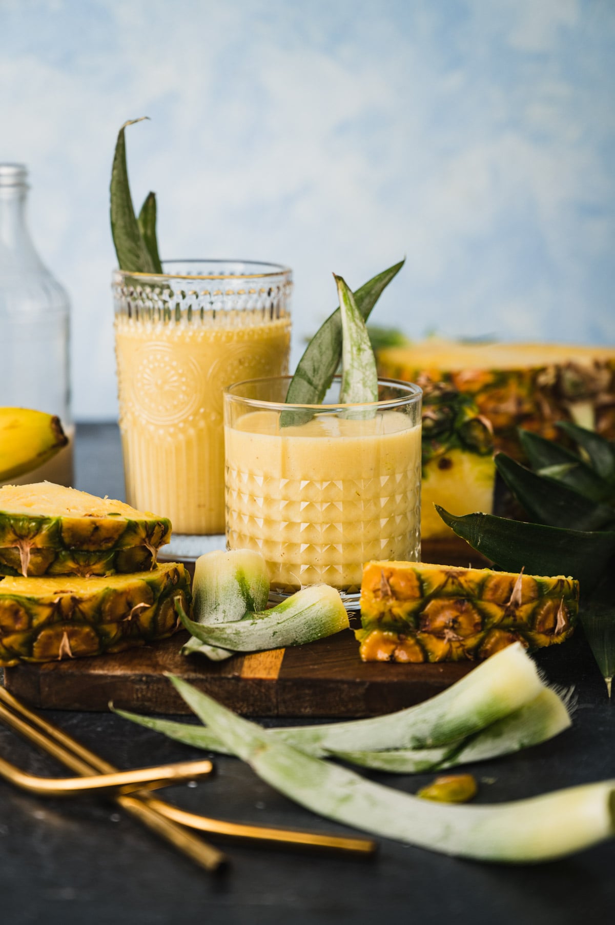 Two glasses of pineapple smoothie are placed on a table along with fresh pineapple slices and leaves. A peeled pineapple and some metal straws are also visible.