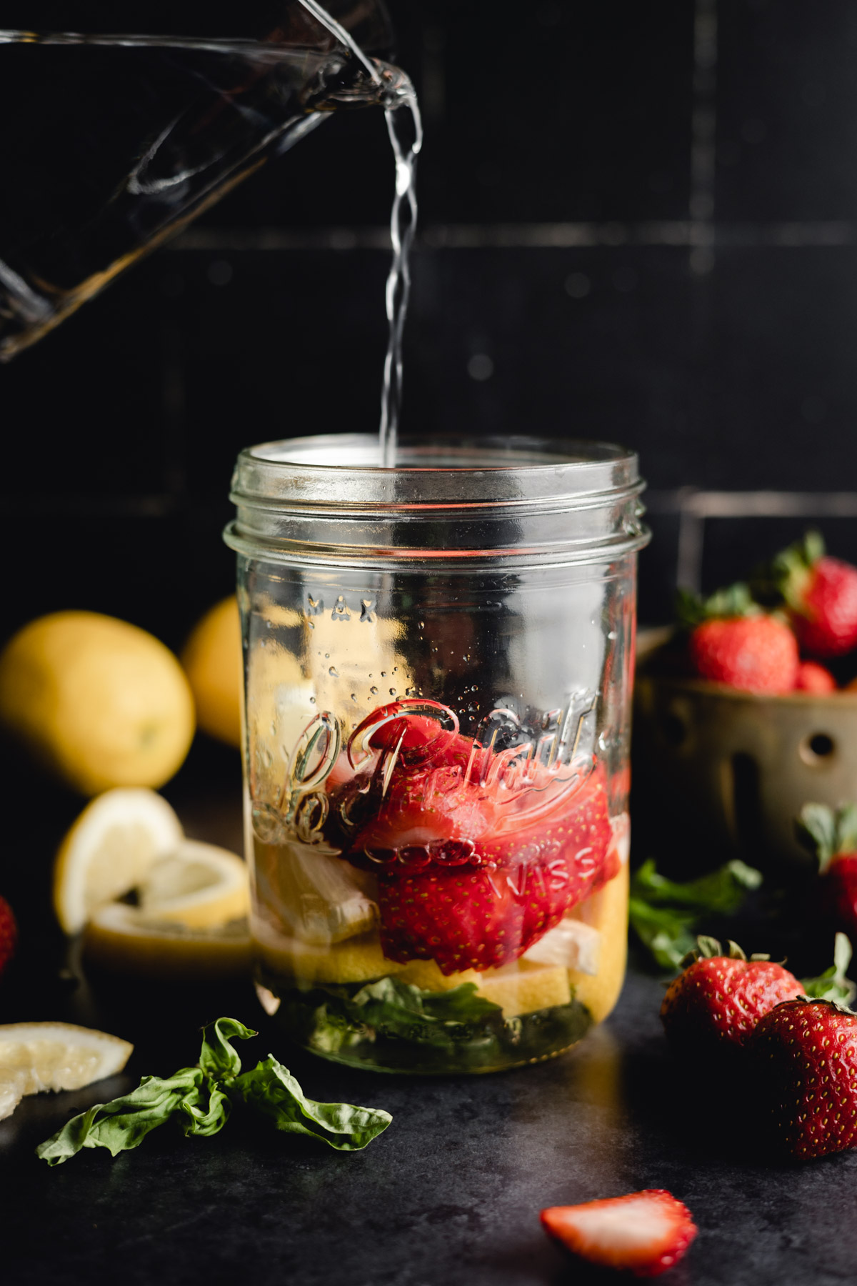 Water being poured into a glass jar containing strawberries, lemon slices, and herbs, with whole strawberries and lemons in the background on a dark surface.