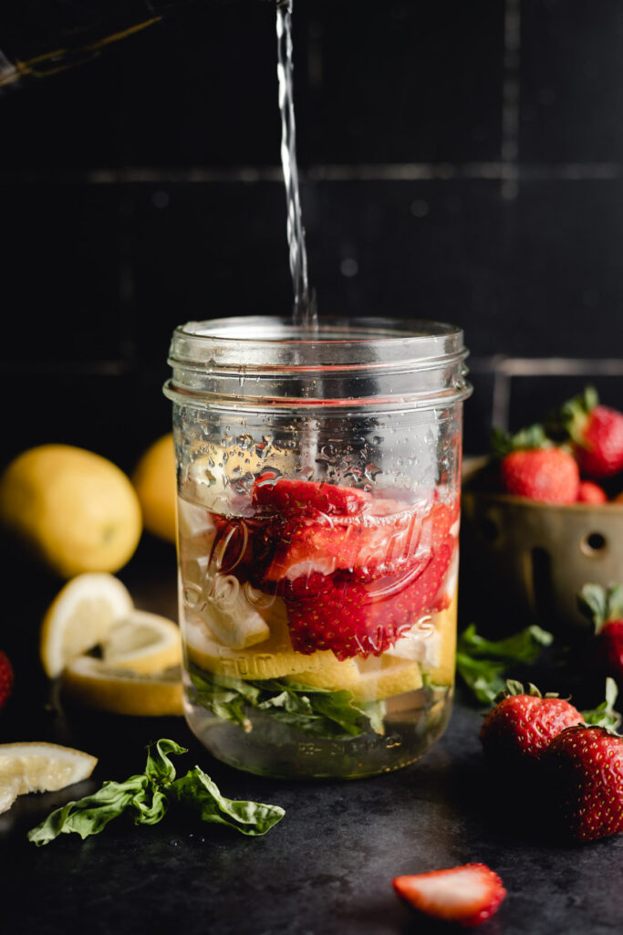 A stream of water is being poured into a mason jar containing sliced strawberries, lemons, and herbs. In the background, there are whole lemons and strawberries.