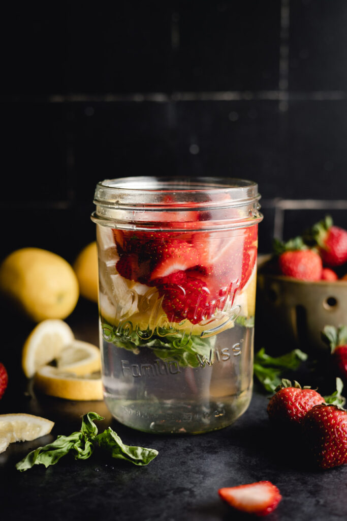 A mason jar filled with water, strawberries, lemon slices, and mint leaves on a dark surface, with additional strawberries, lemon, and mint around it.