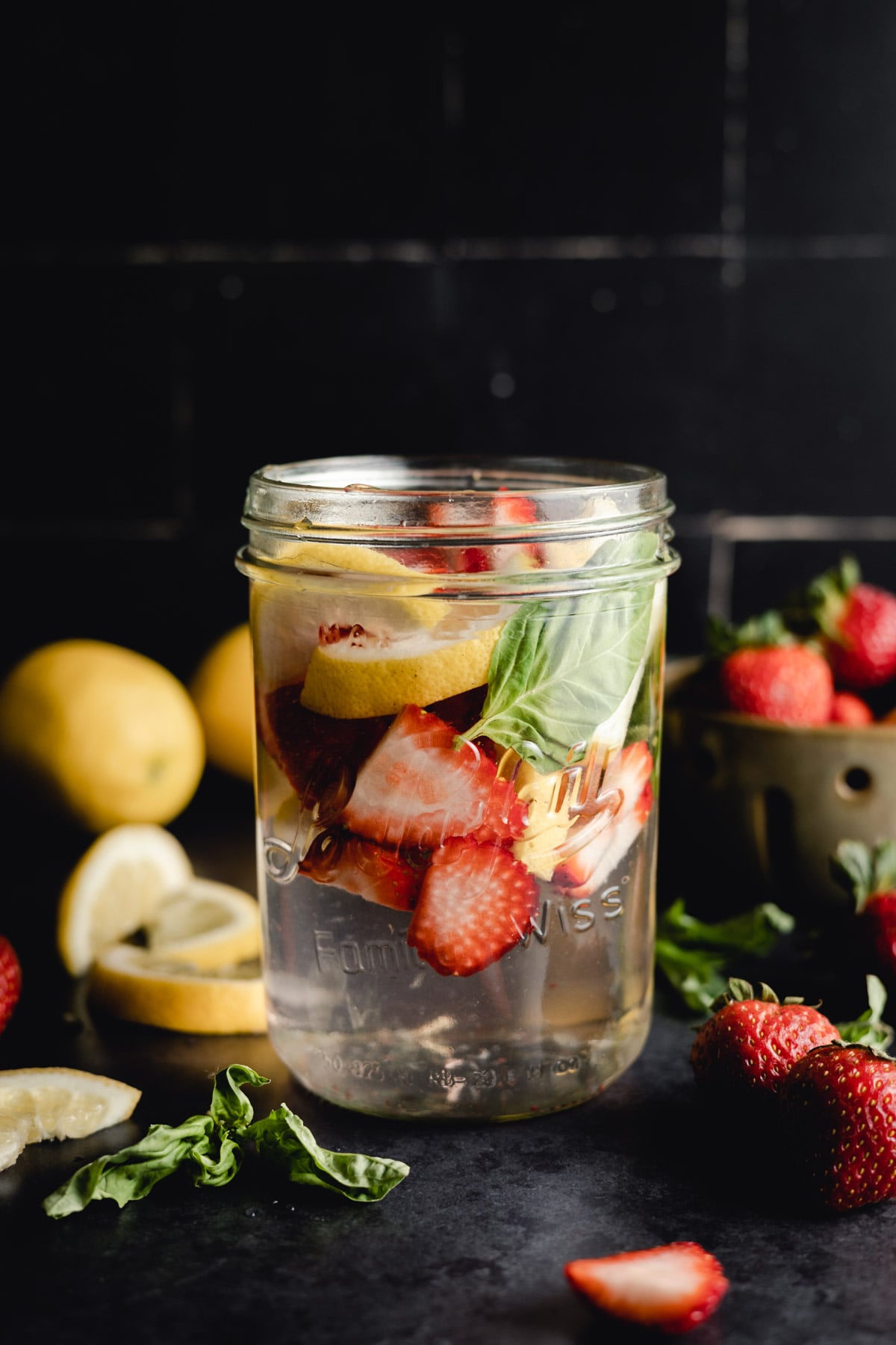 A mason jar filled with water, sliced strawberries, lemon wedges, and basil leaves, surrounded by fresh strawberries, lemons, and scattered basil on a black surface.