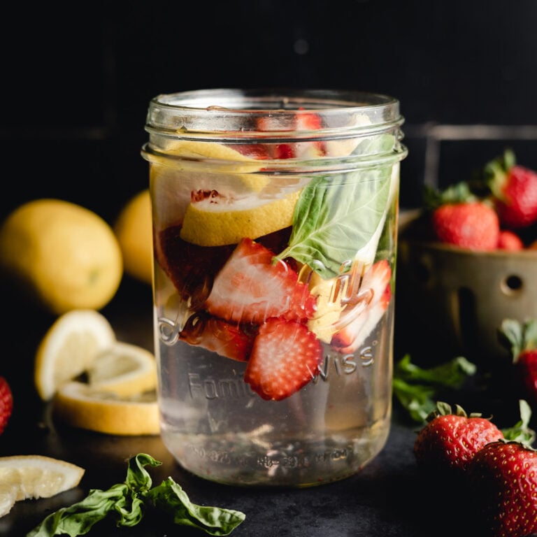 A mason jar filled with water, sliced strawberries, lemon slices, and basil leaves. Fresh strawberries, lemon slices, and basil leaves are scattered around the jar on a dark surface.