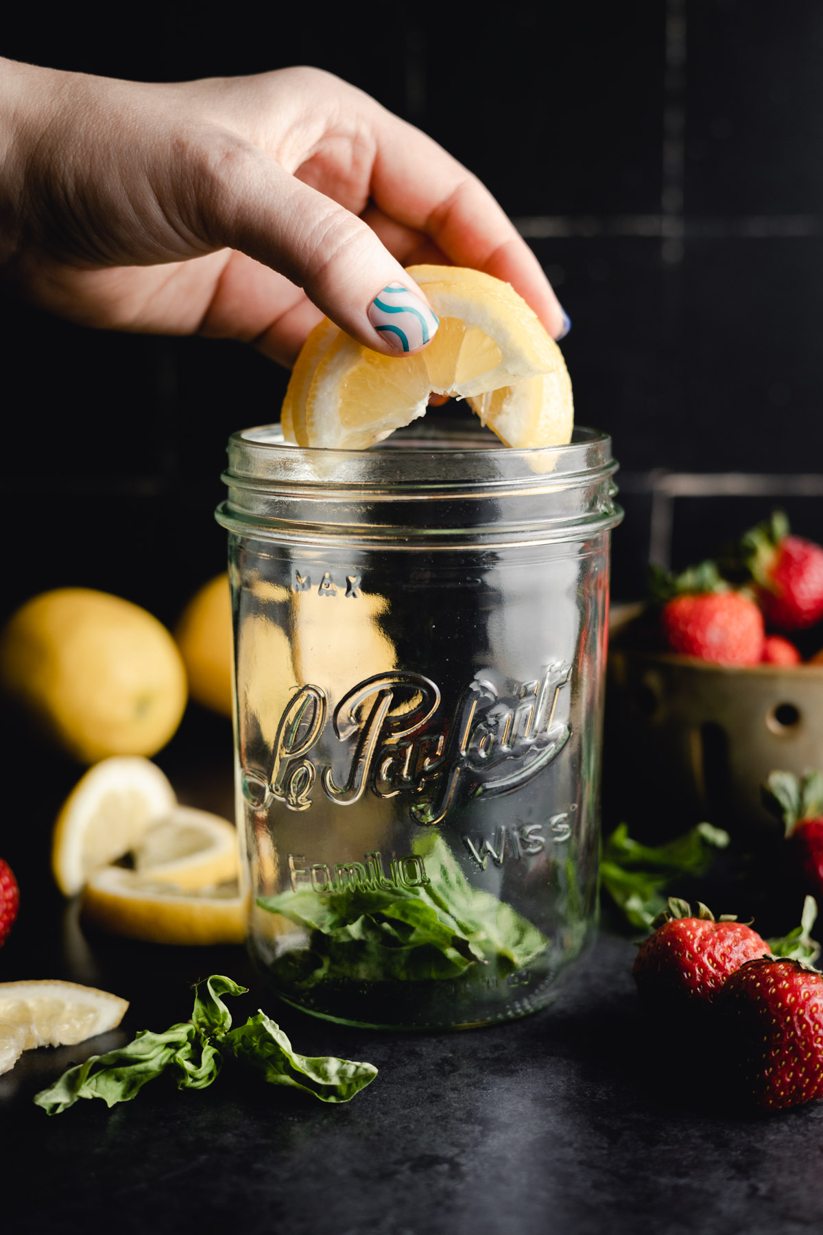 A hand with painted nails places a lemon slice into a glass jar labeled "Le Parfait" among strawberries and greens on a dark surface.