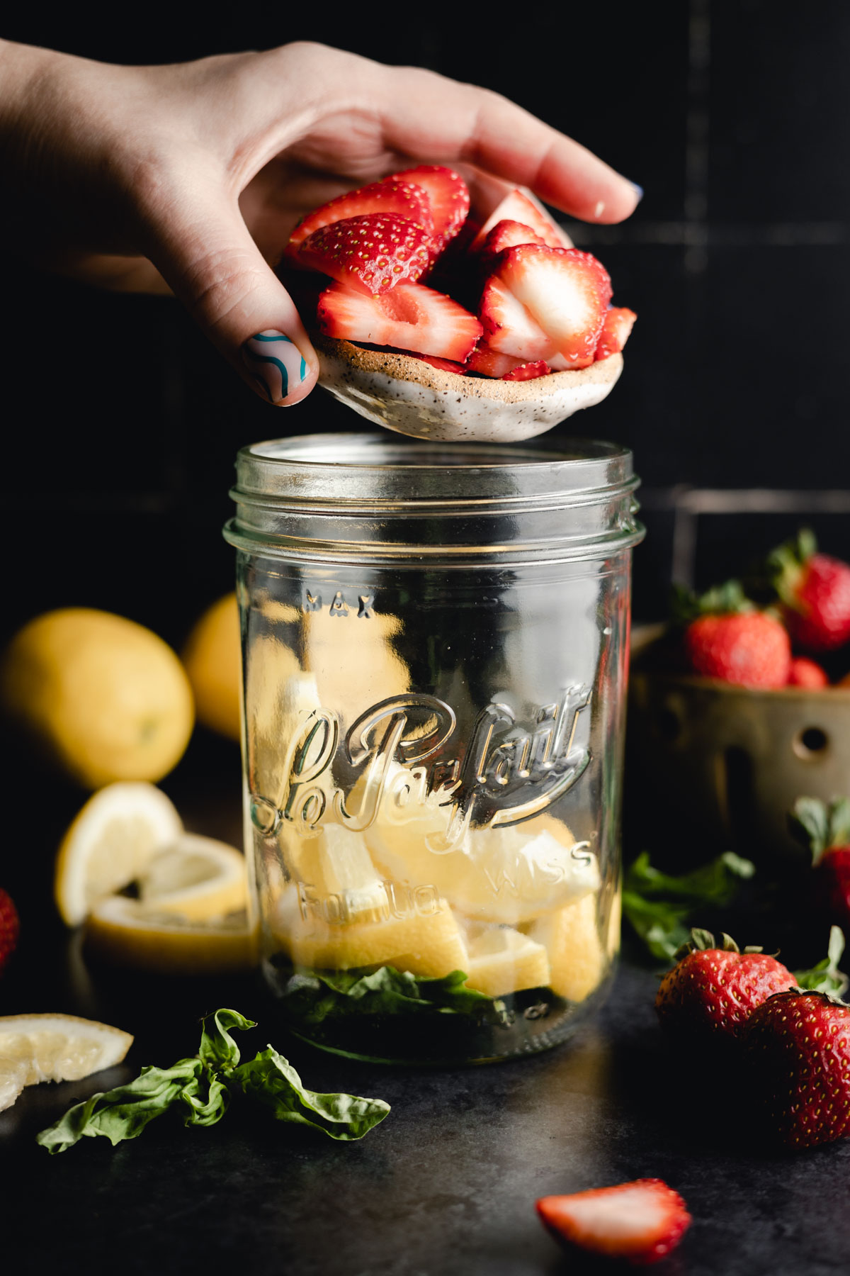 A hand places strawberry slices onto a lemon layer in a glass jar. Fresh strawberries, lemon slices, and leafy greens are scattered around.