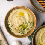 A bowl of creamy hummus topped with chopped herbs, black pepper, and a drizzle of olive oil, placed on a light-colored surface with a wooden utensil and grilled bread partially visible nearby.