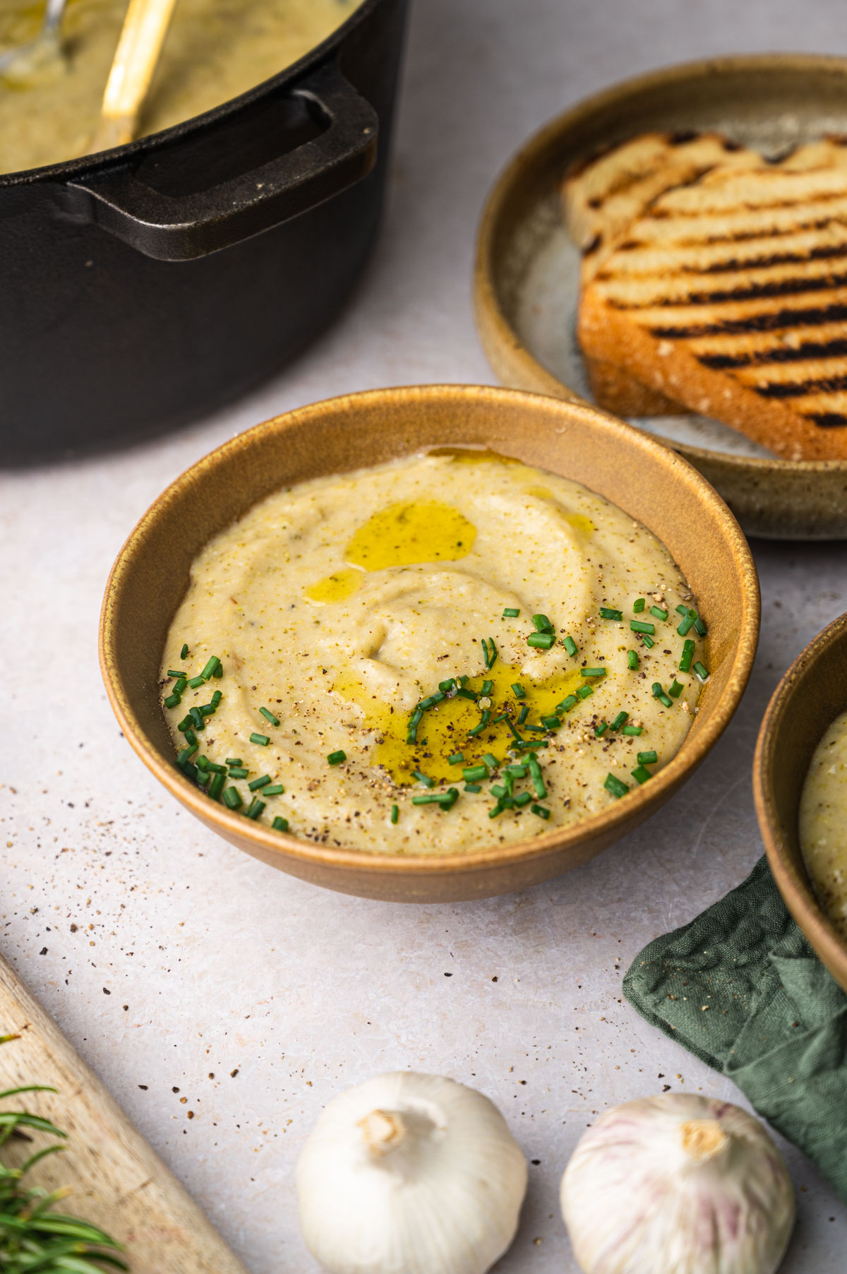 A bowl of creamy soup garnished with chives and olive oil, placed next to a plate of toasted bread. Garlic and a pot of soup are in the background.