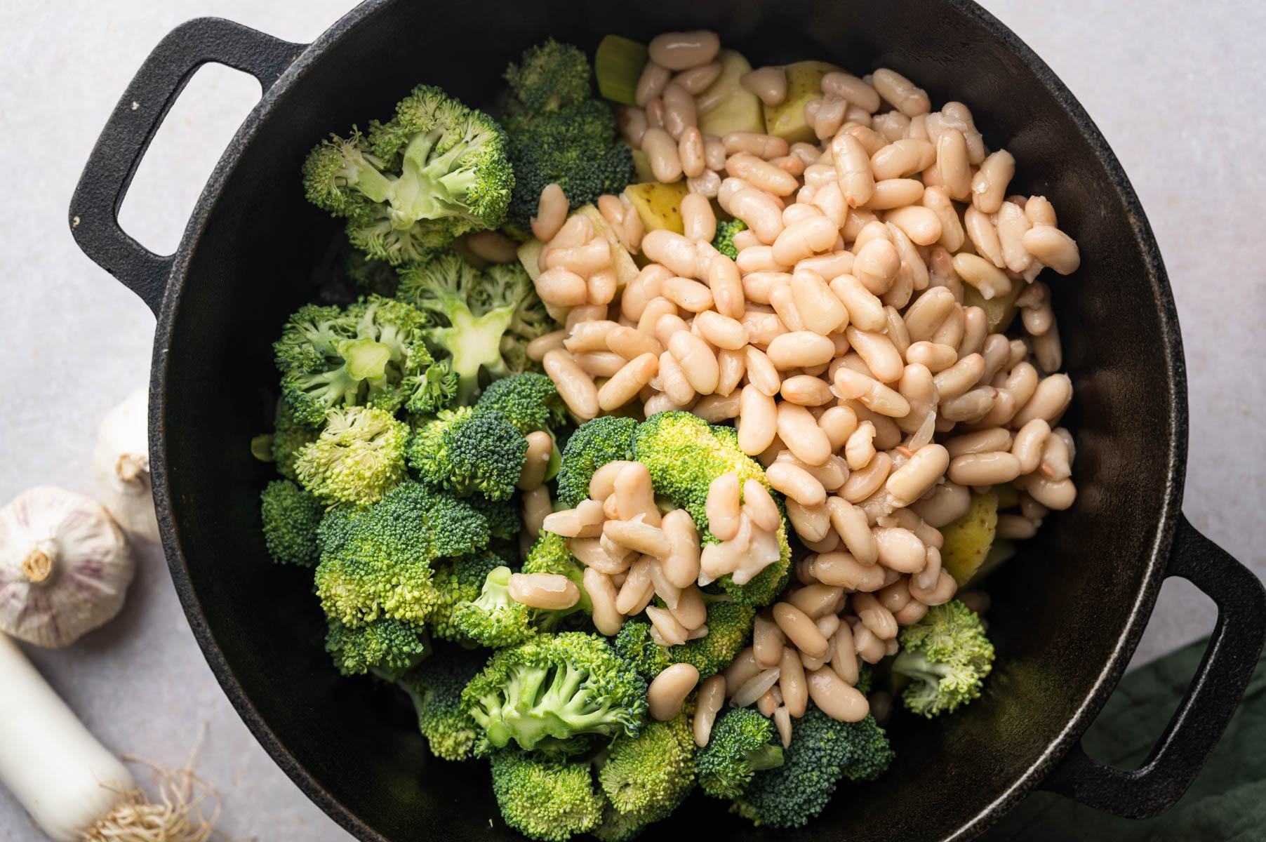 A black cast iron pot filled with chopped broccoli, white beans, and sliced potatoes. Garlic bulbs and a leek are on the side.