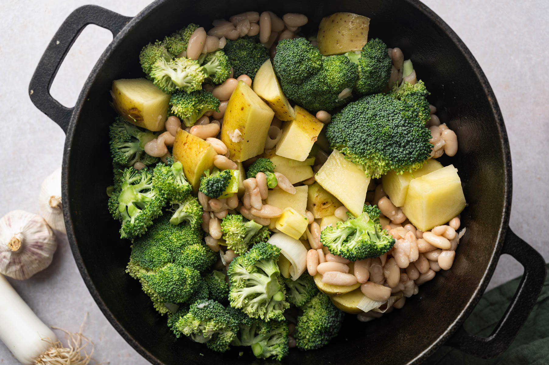 A black pot filled with broccoli florets, diced potatoes, and white beans. Garlic and leeks are visible in the background on the countertop.