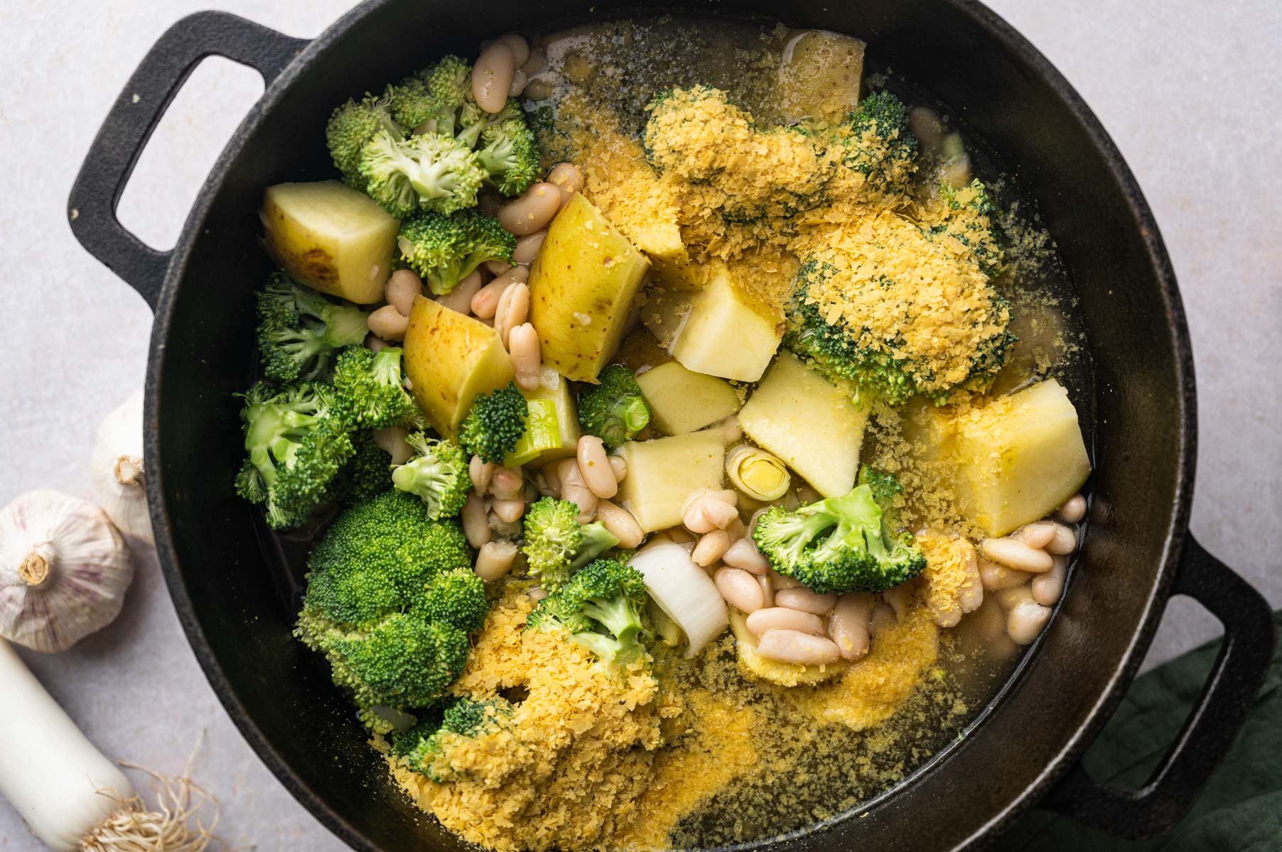 A cast iron pot filled with broccoli, potatoes, white beans, and a yellow seasoning, partially covered with a liquid. Garlic cloves and a bulb, and a leek are partially visible beside the pot.