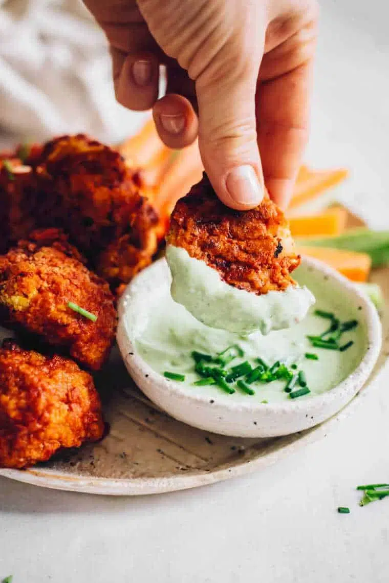 A person dips a crispy falafel ball into a small bowl of green sauce, garnished with chopped chives, with more falafel and tasty dishes in the background.