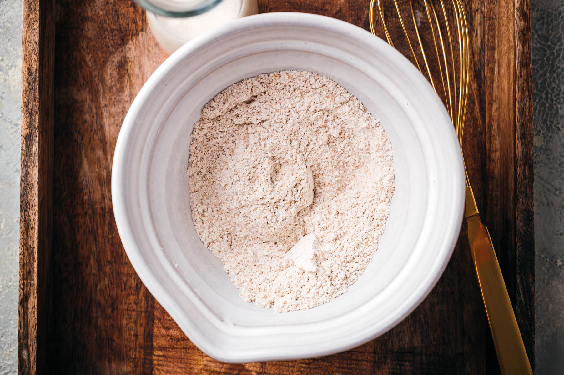 A white ceramic bowl filled with flour sits on a wooden surface next to a gold-colored whisk.