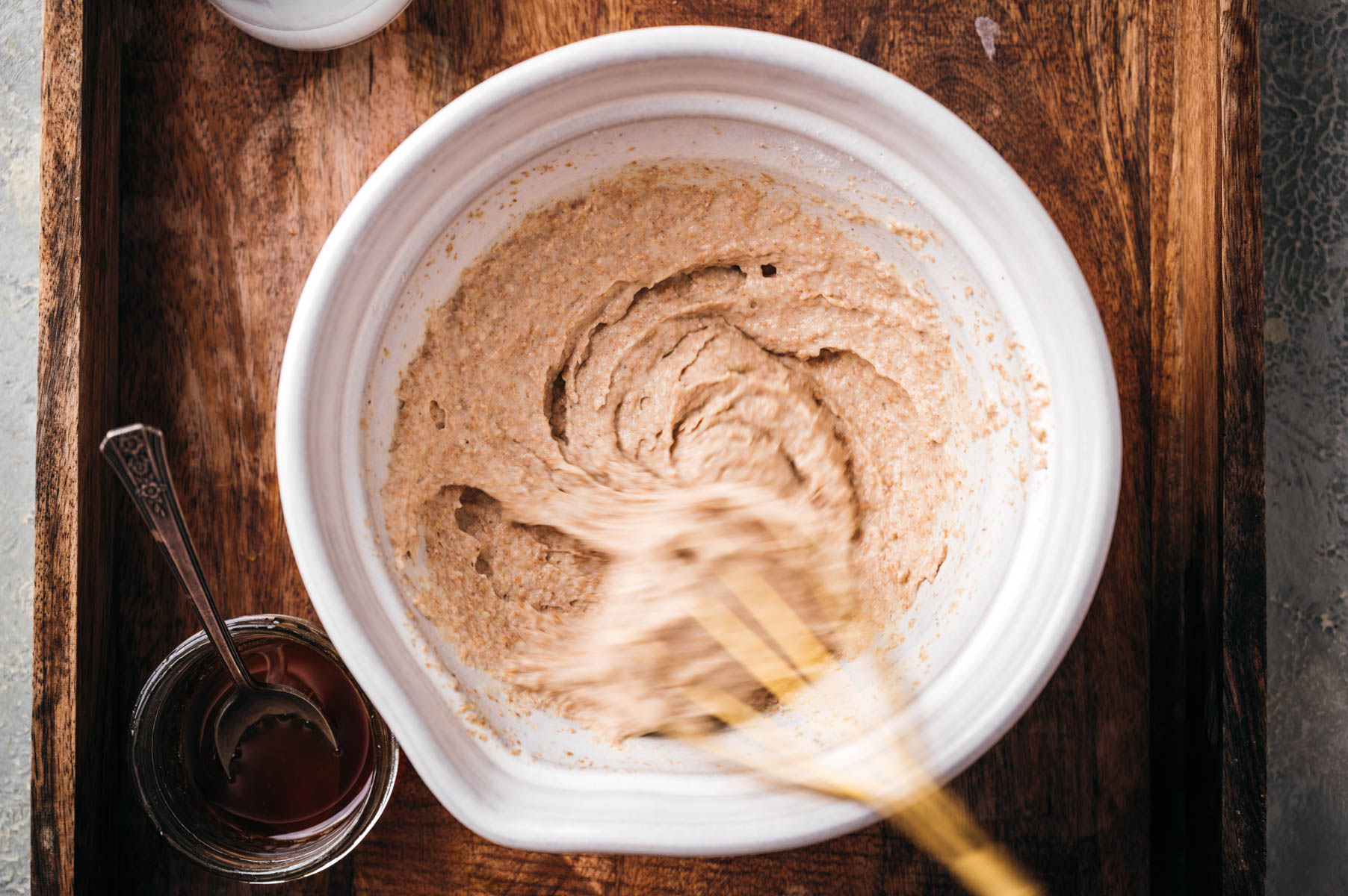A wooden spoon stirring batter in a white bowl on a wooden surface, beside is a small jar of honey with a spoon in it.