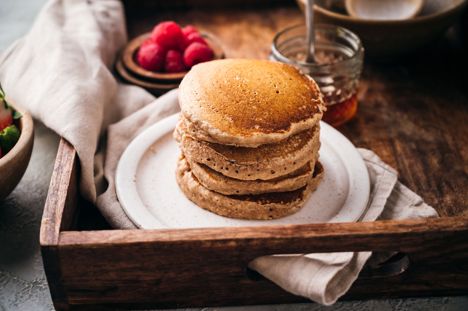 A stack of pancakes on a white plate inside a wooden tray, accompanied by a bowl of raspberries and a jar of syrup.