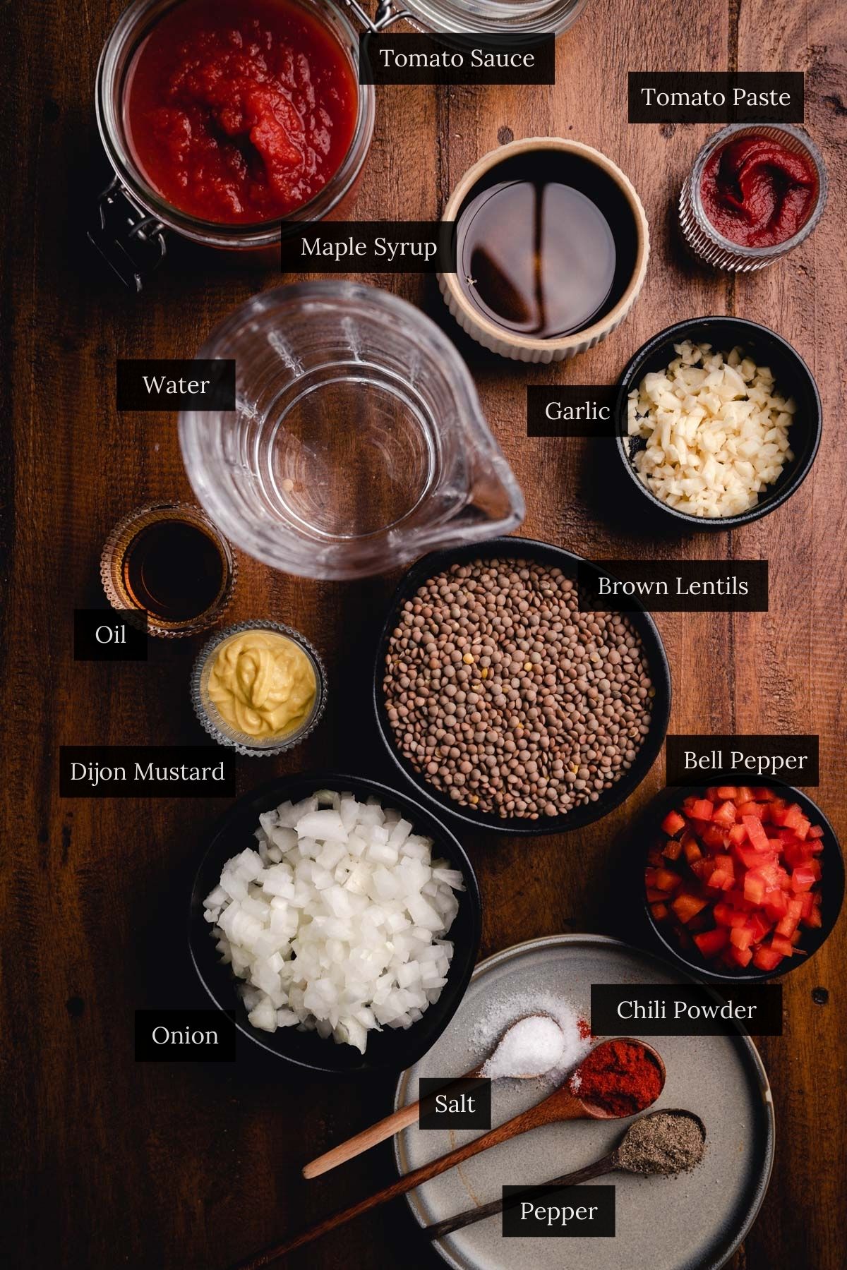 Top-down view of various ingredients for vegan sloppy joes labeled on a wooden surface, including tomato sauce, water, garlic, brown lentils, bell pepper, onion, chili powder, and more.