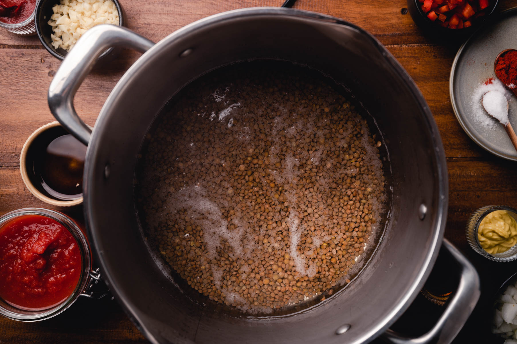 Lentils boiling in a pot, surrounded by ingredients like rice, spices, and tomato sauce on a wooden table.
