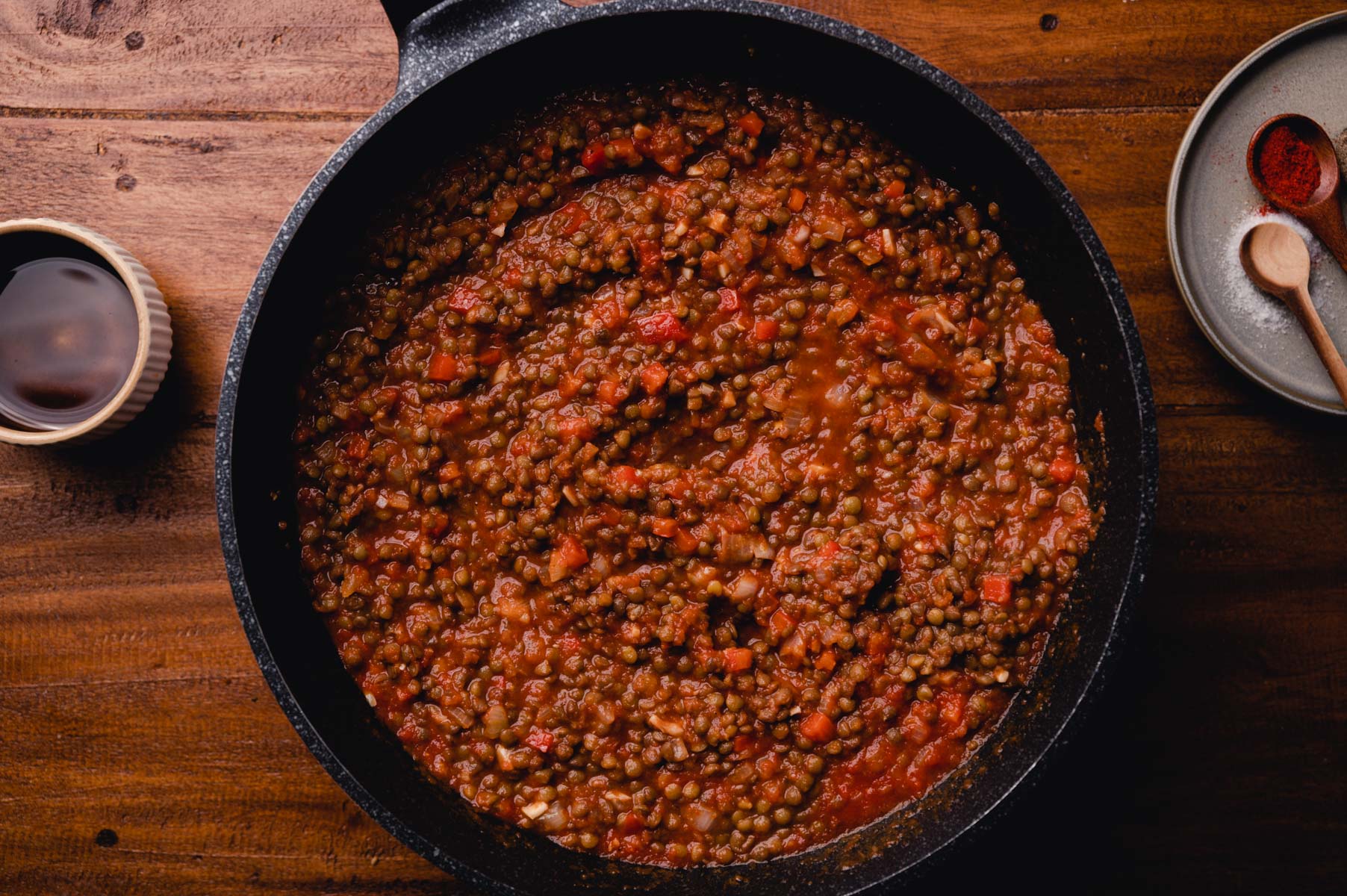 A skillet of cooked lentils and tomatoes on a wooden surface, accompanied by spices and a small bowl of sauce.