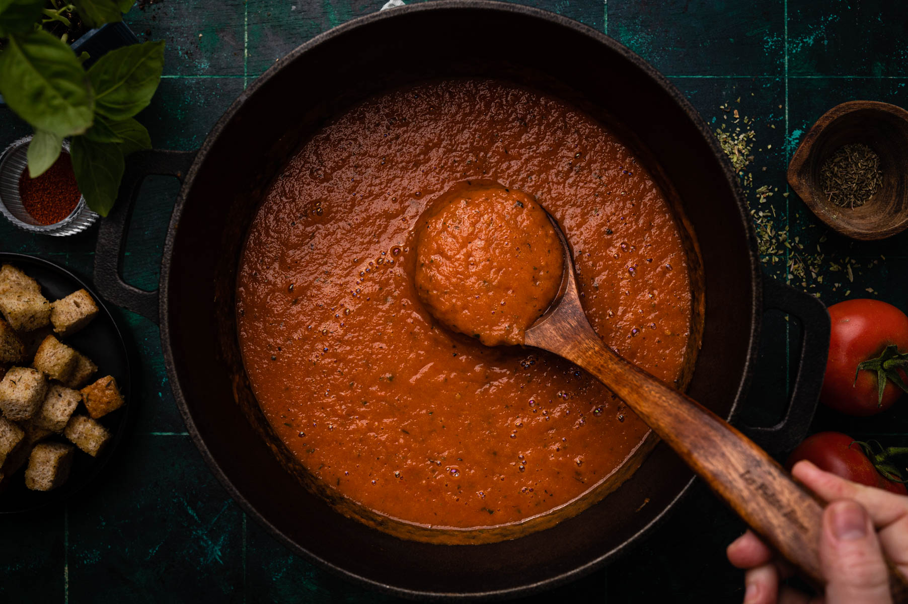 A wooden spoon scooping rich tomato sauce from a cast iron skillet, surrounded by fresh basil, spices, and croutons on a dark tabletop.