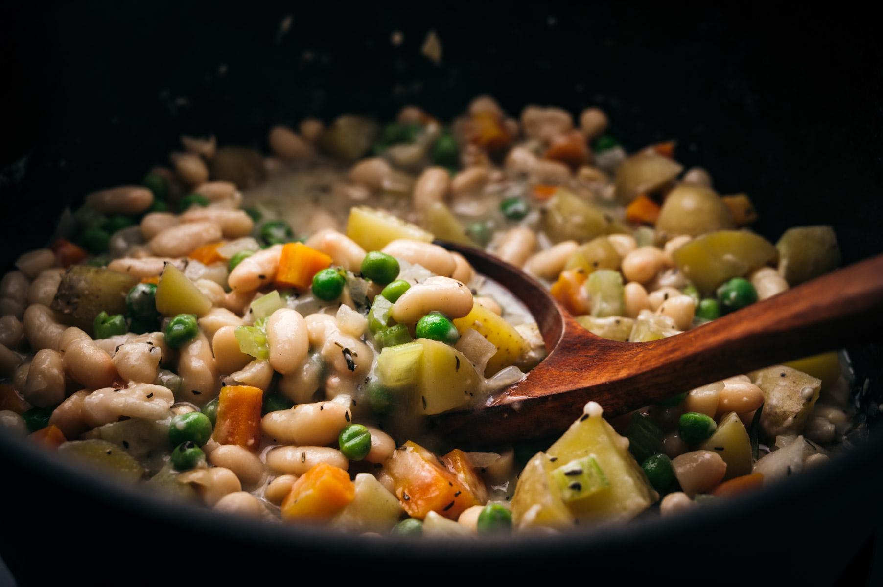 A close-up of a vegetable stew with beans, peas, carrots, and potatoes in a black pot, stirred with a wooden spoon.