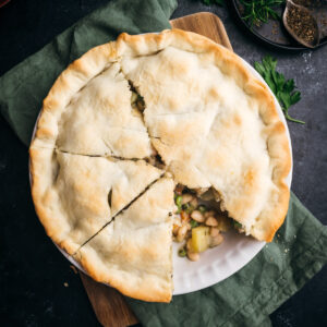 A homemade pot pie with a flaky crust, partially sliced, revealing a savory filling with vegetables and beans, served on a wooden board.