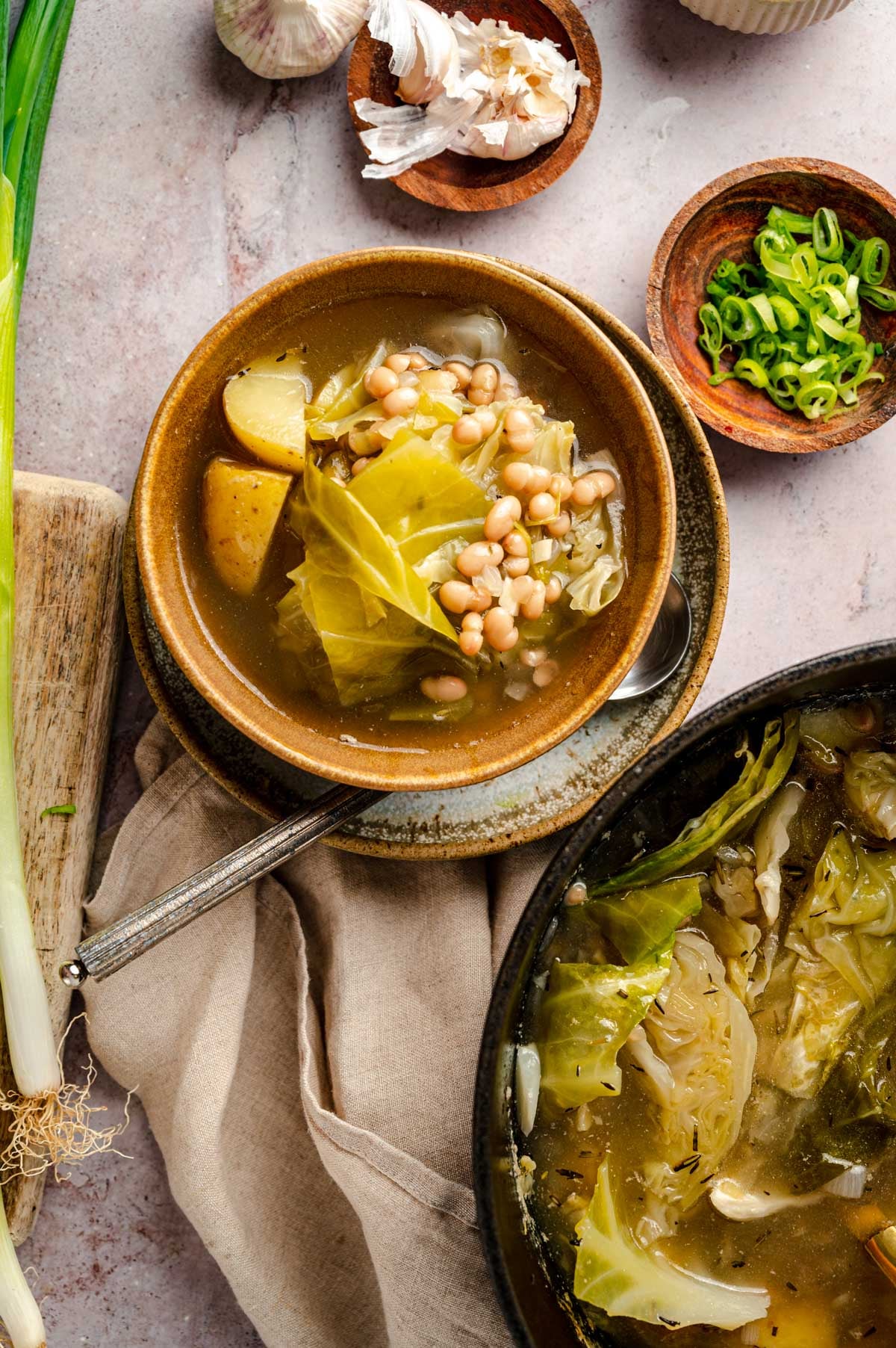 A bowl of vegetable soup with potatoes, cabbage, and white beans next to a plate of chopped green onions and garlic cloves. Another pot of soup is partially visible in the bottom right.