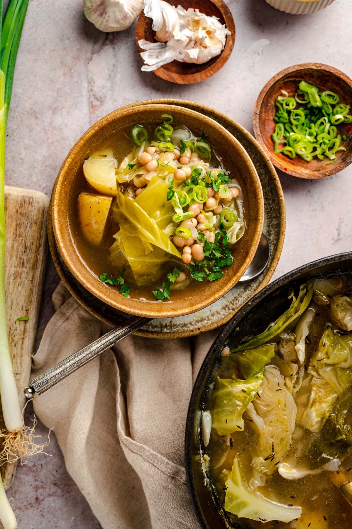 A bowl of vegetable soup with cabbage, potatoes, white beans, and green onions next to a pot of the same soup. Garlic and chopped green onions are in small dishes beside the bowl.