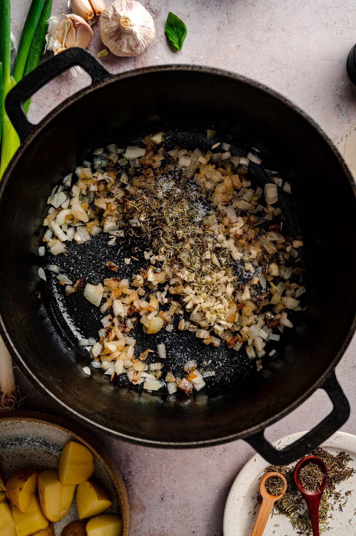 A cast iron pot on a countertop contains sautéed onions and seasoning. Surrounding the pot are garlic, green onions, a small bowl of diced potatoes, and measuring spoons with spices.
