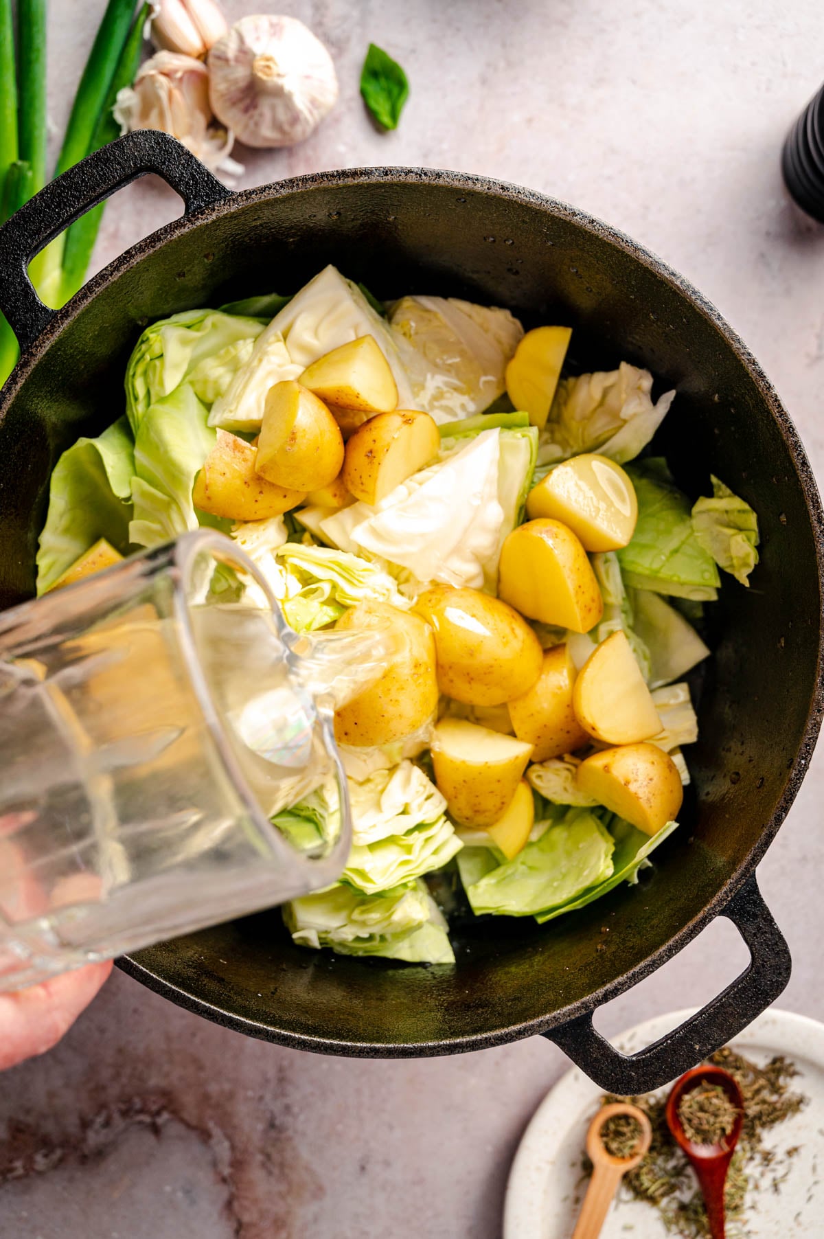 A pot containing chopped cabbage and quartered potatoes has water being poured into it. Garlic, green onions, and seasonings are visible on the sides.