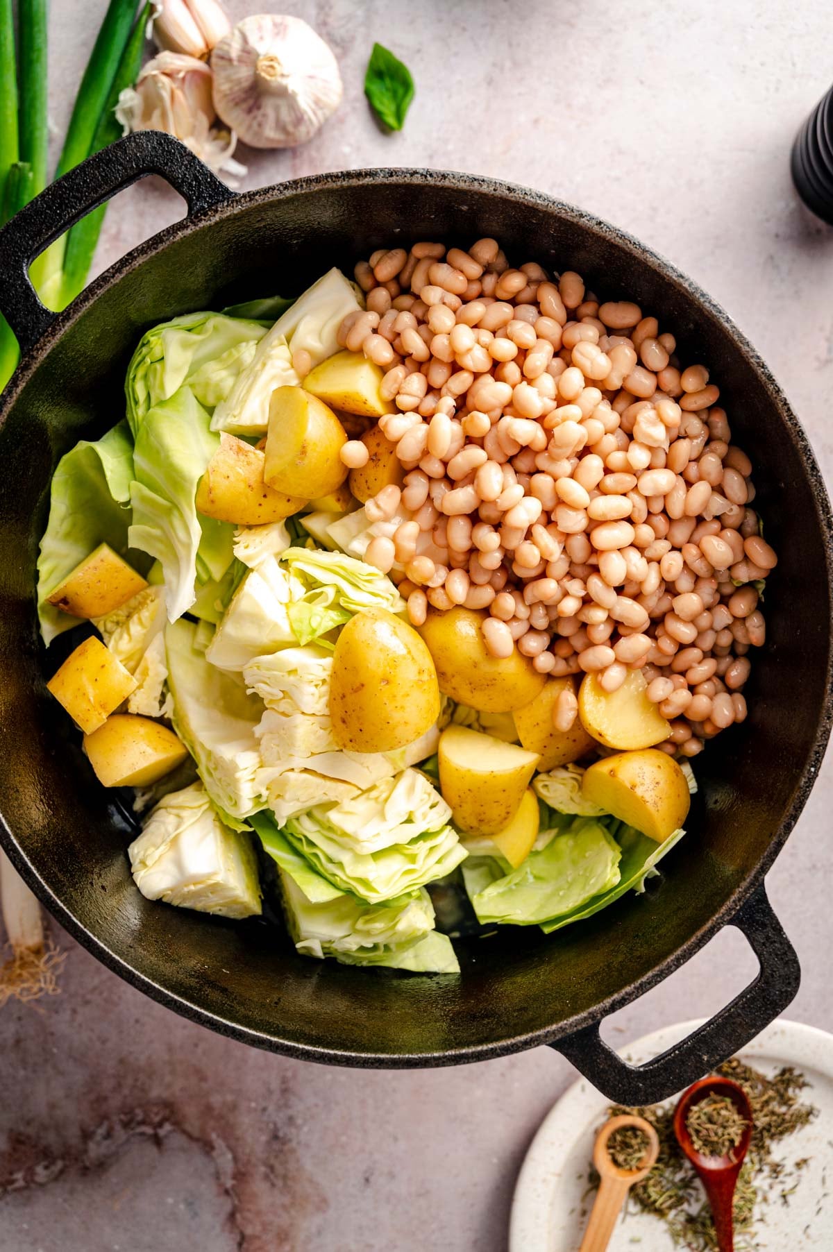 A cast iron pot contains chopped cabbage, white beans, and diced yellow potatoes, placed on a light background. Garlic bulbs, green onions, and a dish of herbs appear nearby.