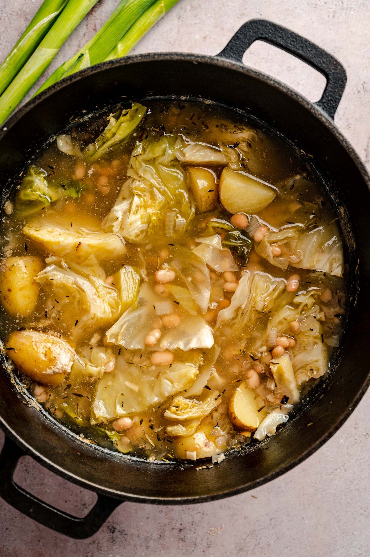 A pot of vegetable soup containing cabbage, potatoes, chickpeas, and broth. Green onion stalks are placed beside the pot.