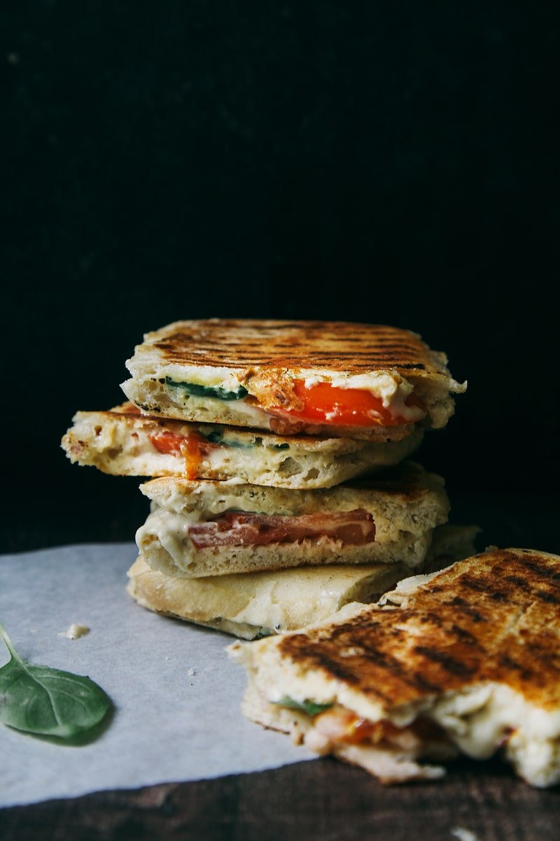 Close-up of a stack of grilled vegetable panini sandwiches with visible layers of tomato, cheese, and spinach on parchment paper against a dark background. Perfect for those who love tasty recipes, these paninis can be ideally served with a warm bowl of butternut squash soup.