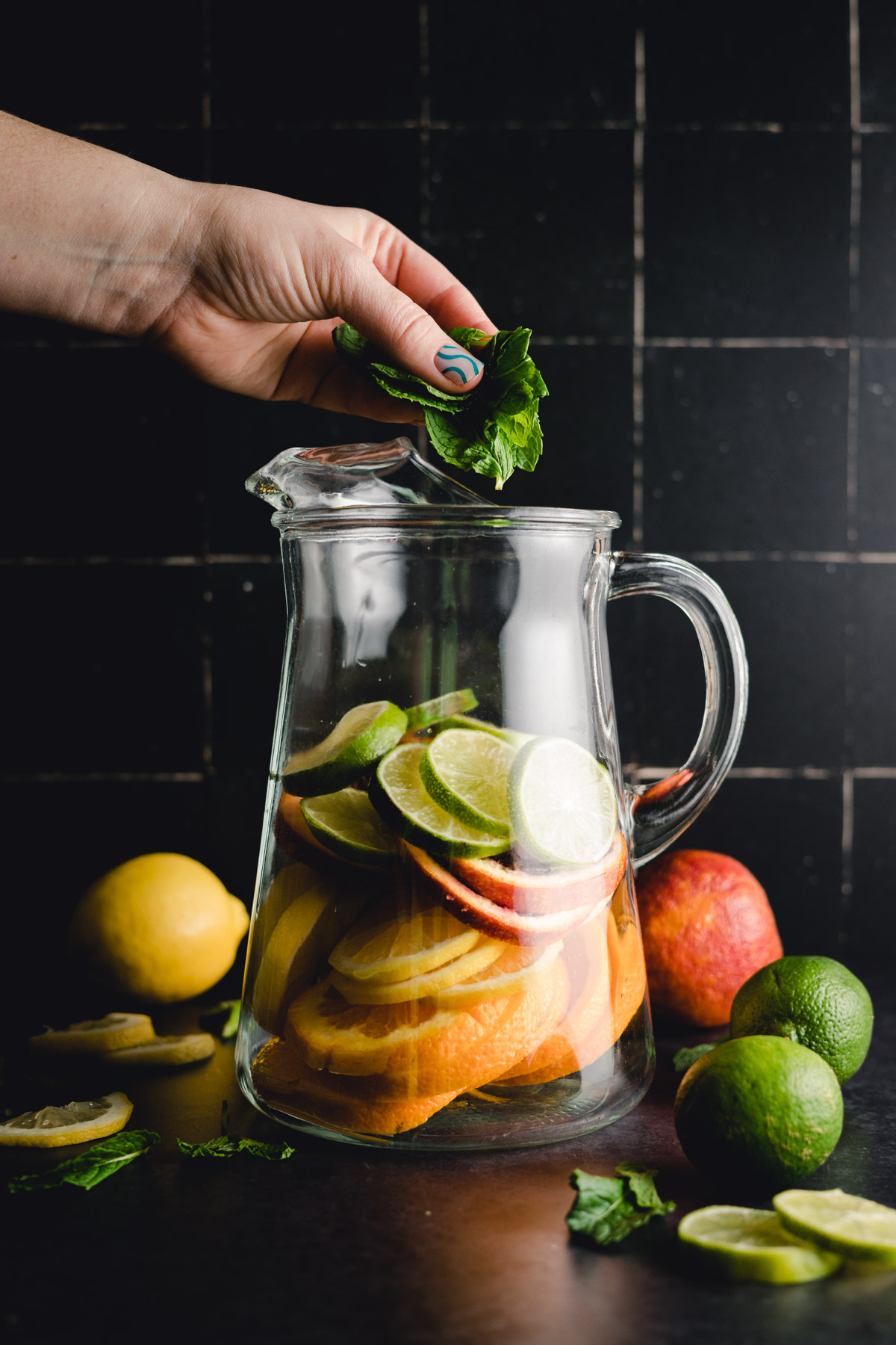 Hand adding mint leaves to a glass pitcher filled with sliced citrus fruits including oranges, lemons, and limes. Whole fruits are placed around the pitcher.