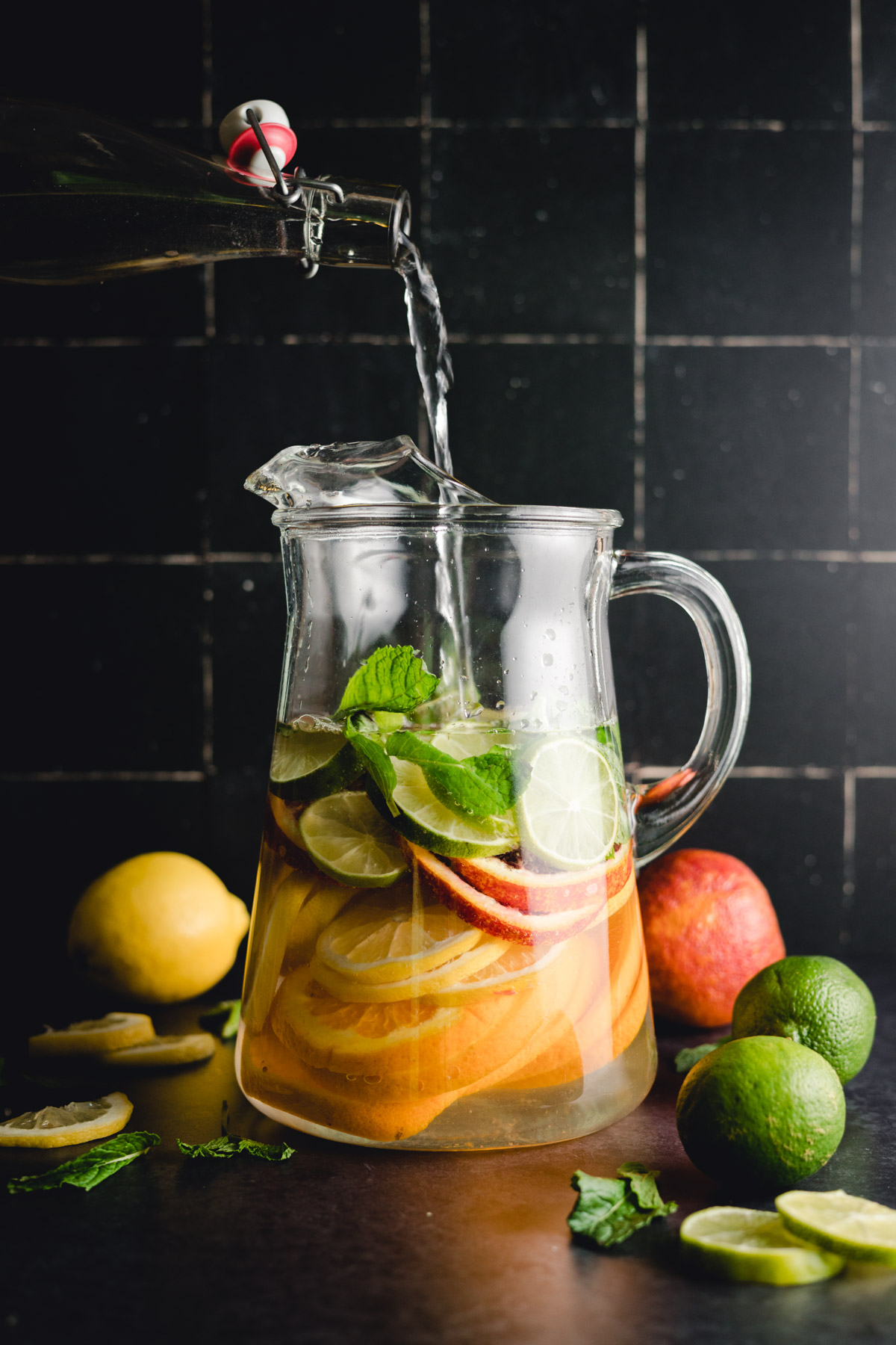 Water being poured into a glass pitcher filled with sliced citrus fruits, mint leaves, and ice. Whole fruits are placed around the pitcher against a dark tiled background.