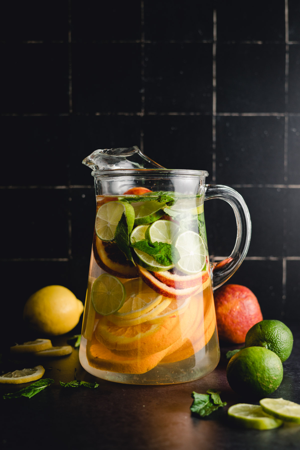 A glass pitcher filled with water, lime, orange, and mint slices. Fresh lemons, limes, a whole orange, and mint leaves are placed around the pitcher on a dark surface.
